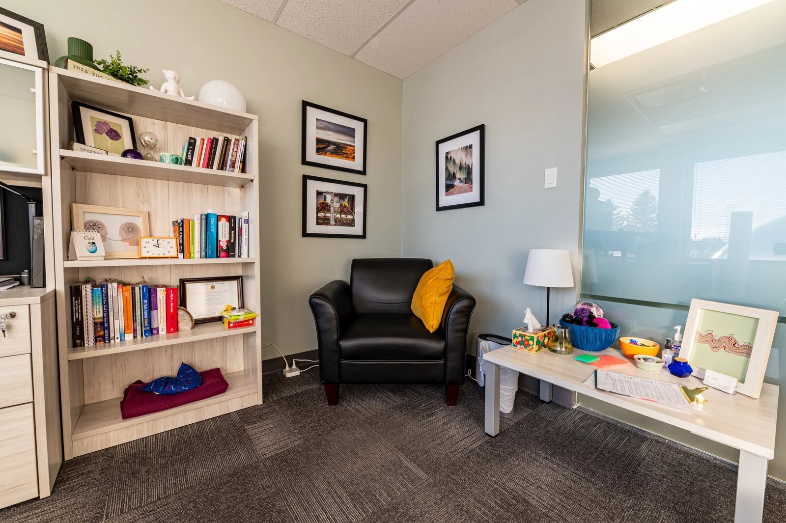 Office corner with a black armchair and yellow pillow, a white table with personal items, framed artwork on the walls, a bookshelf with books and decorative items, and a large window with blinds.