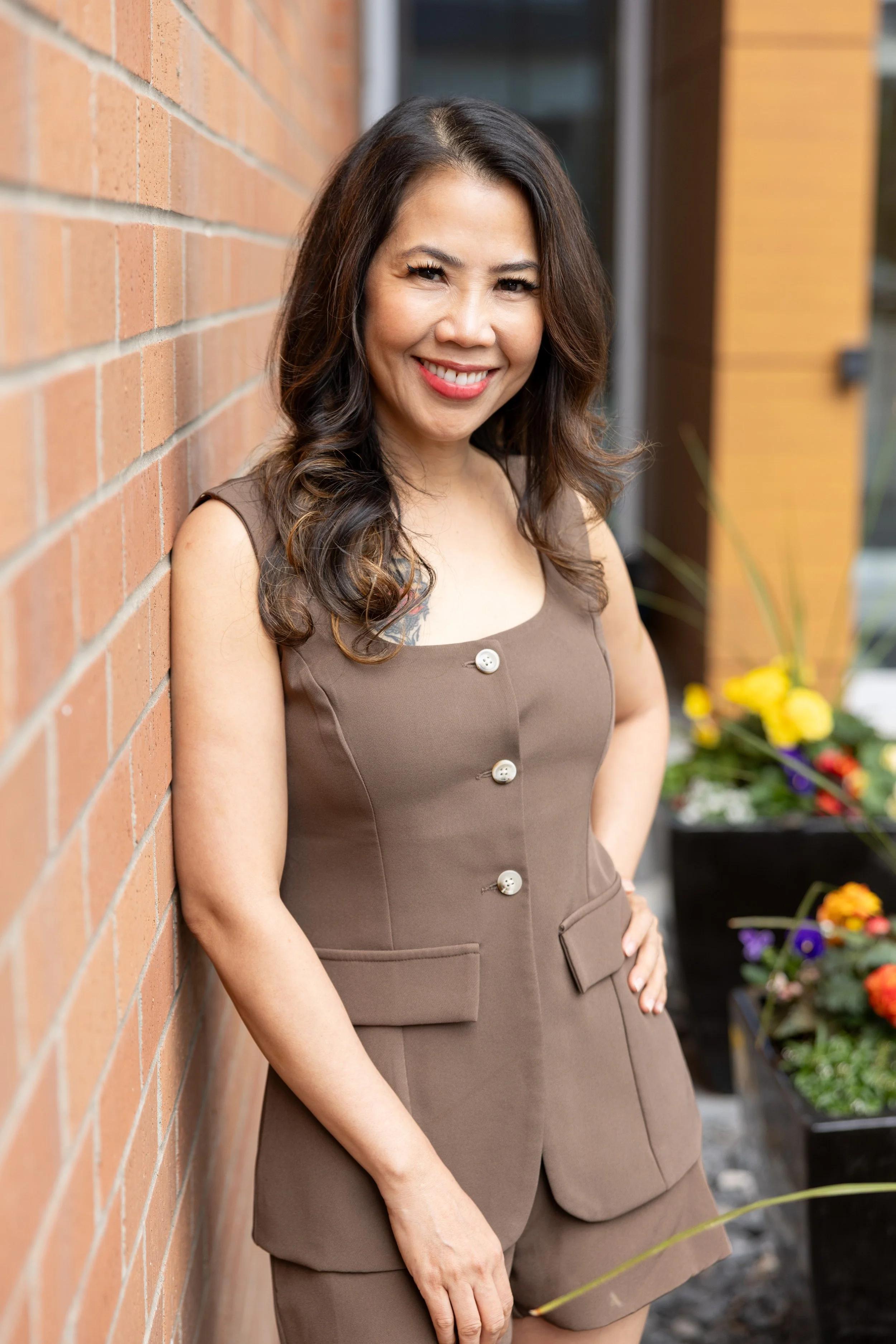A woman with dark hair and a smile, wearing a brown dress with large buttons, standing outdoors next to a brick wall with colorful flowers in the background.
