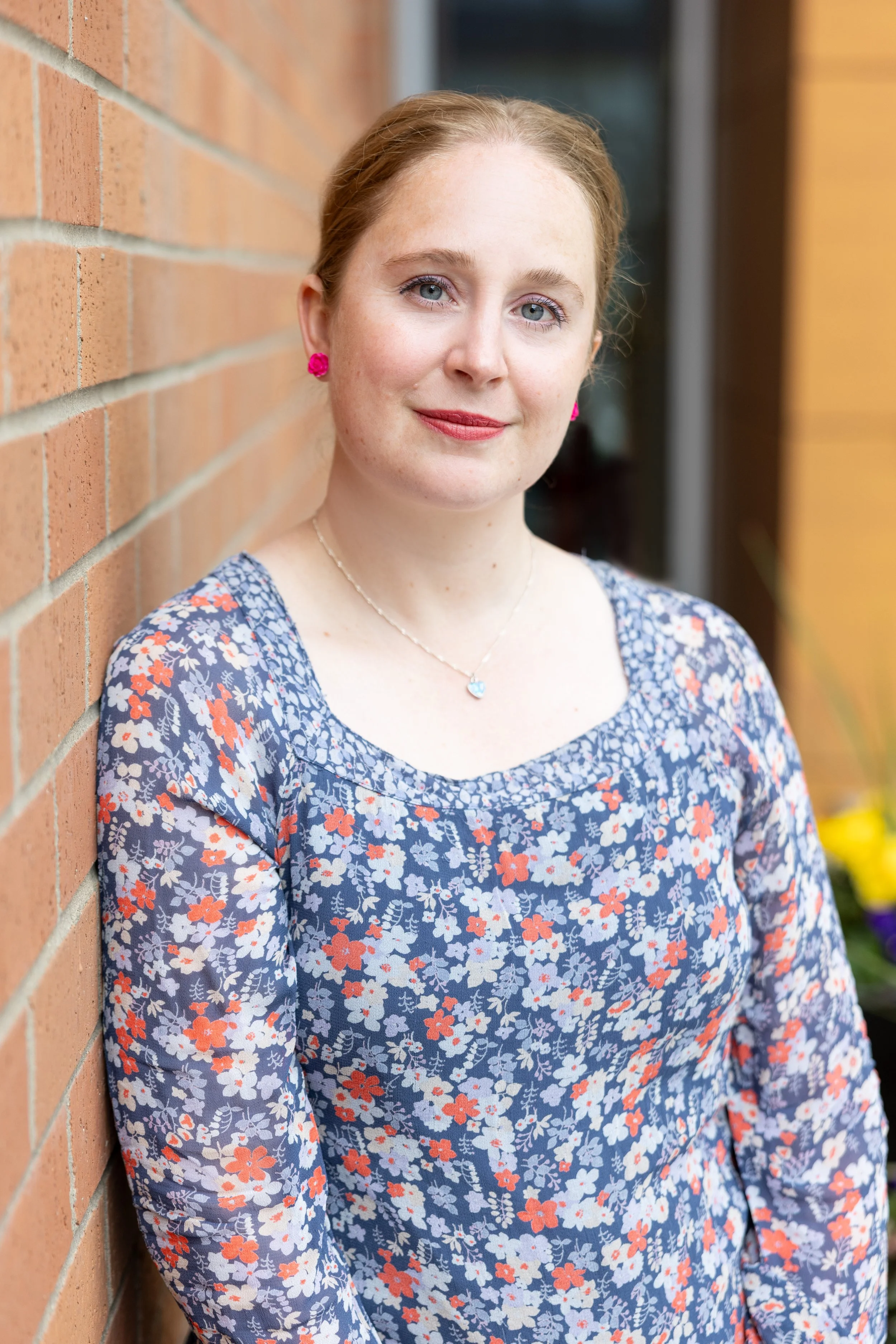 A woman with red hair and blue eyes leaning against a brick wall, wearing a floral dress with shades of blue, white, and red, pink earrings, a necklace with a blue heart pendant, and light makeup with lipstick.