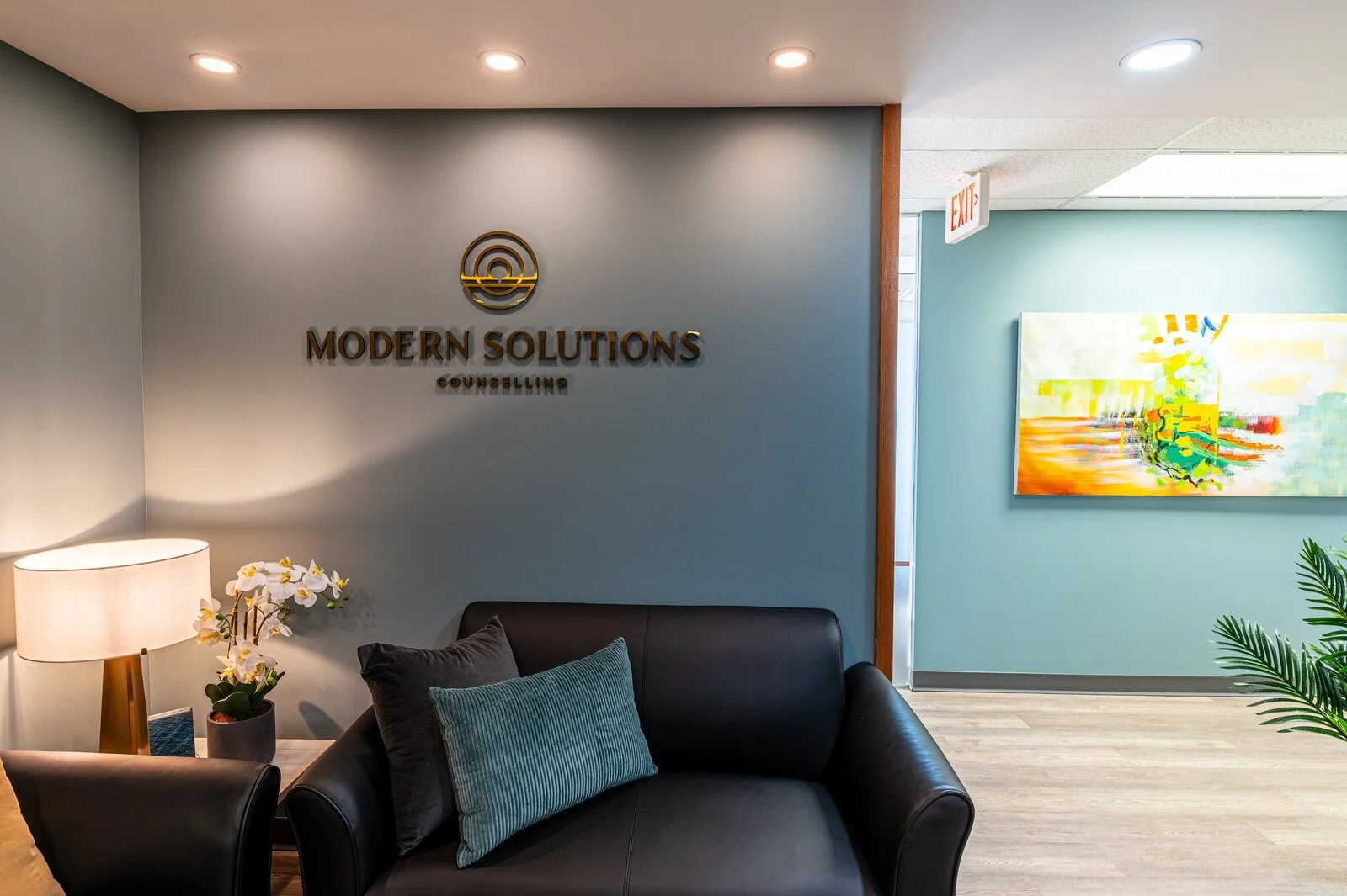 Reception area with a sign that reads 'Modern Solutions Counseling' on the wall, a sofa with cushions, a lamp, and a flowerpot with white orchids. There is a colorful abstract painting on the adjacent wall, and an exit sign near the ceiling.