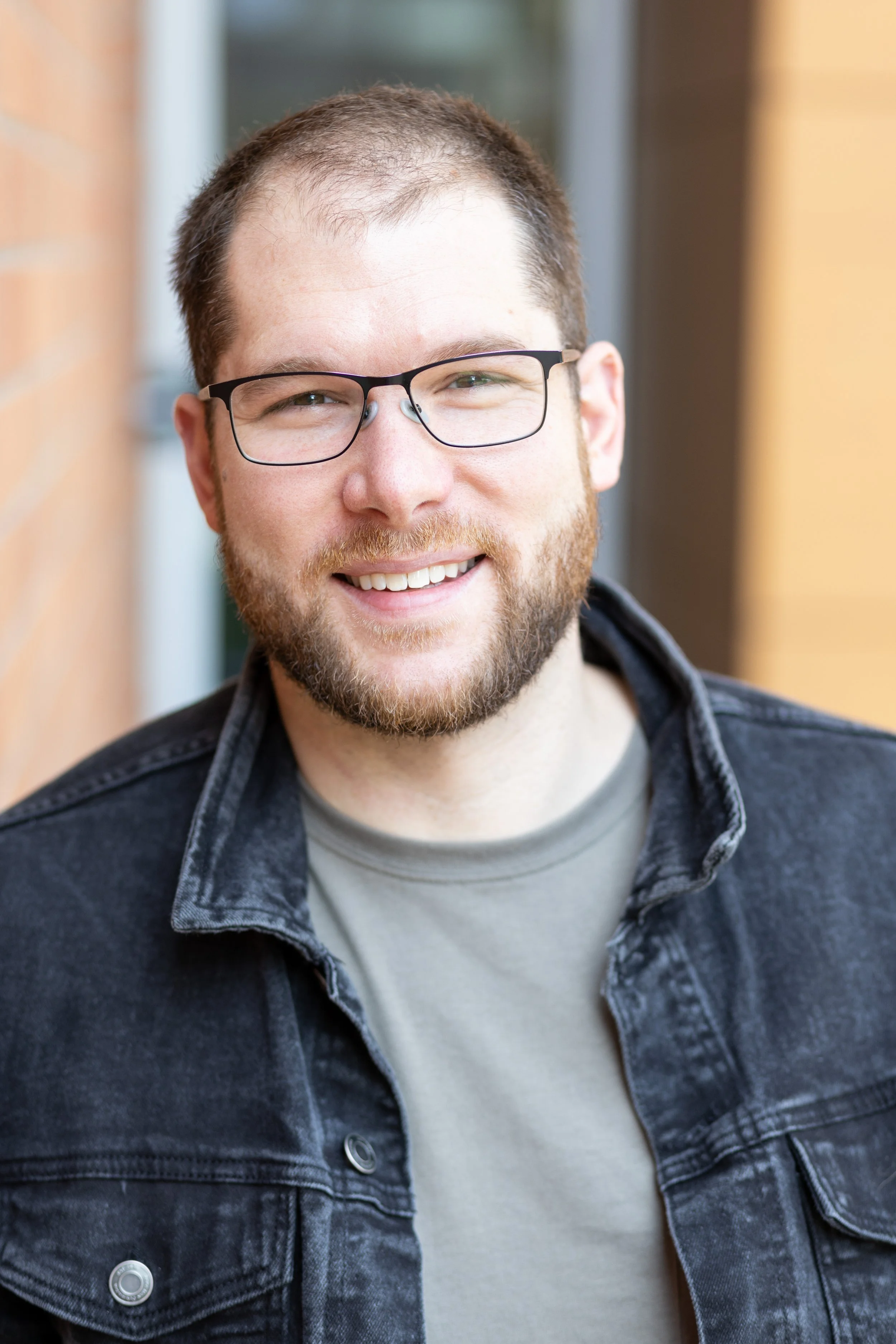Close-up of a smiling man with glasses, a beard, and short hair, wearing a dark denim jacket and a light-colored shirt, standing outdoors near a brick wall with blurred background.