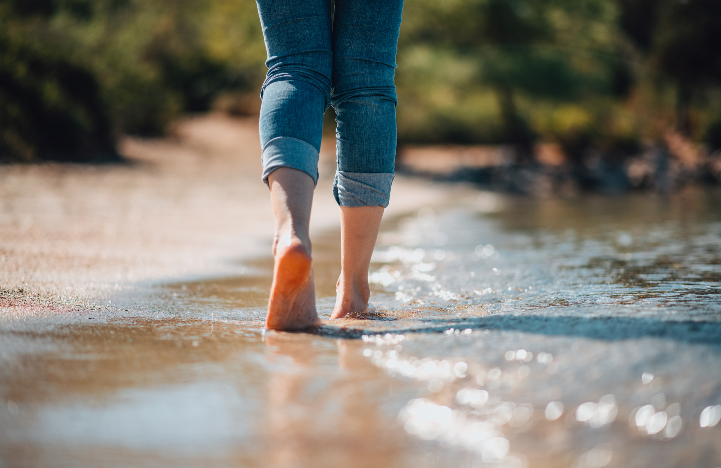Person walking barefoot along the shoreline at a beach, with rolled-up jeans and water splashing around their feet.