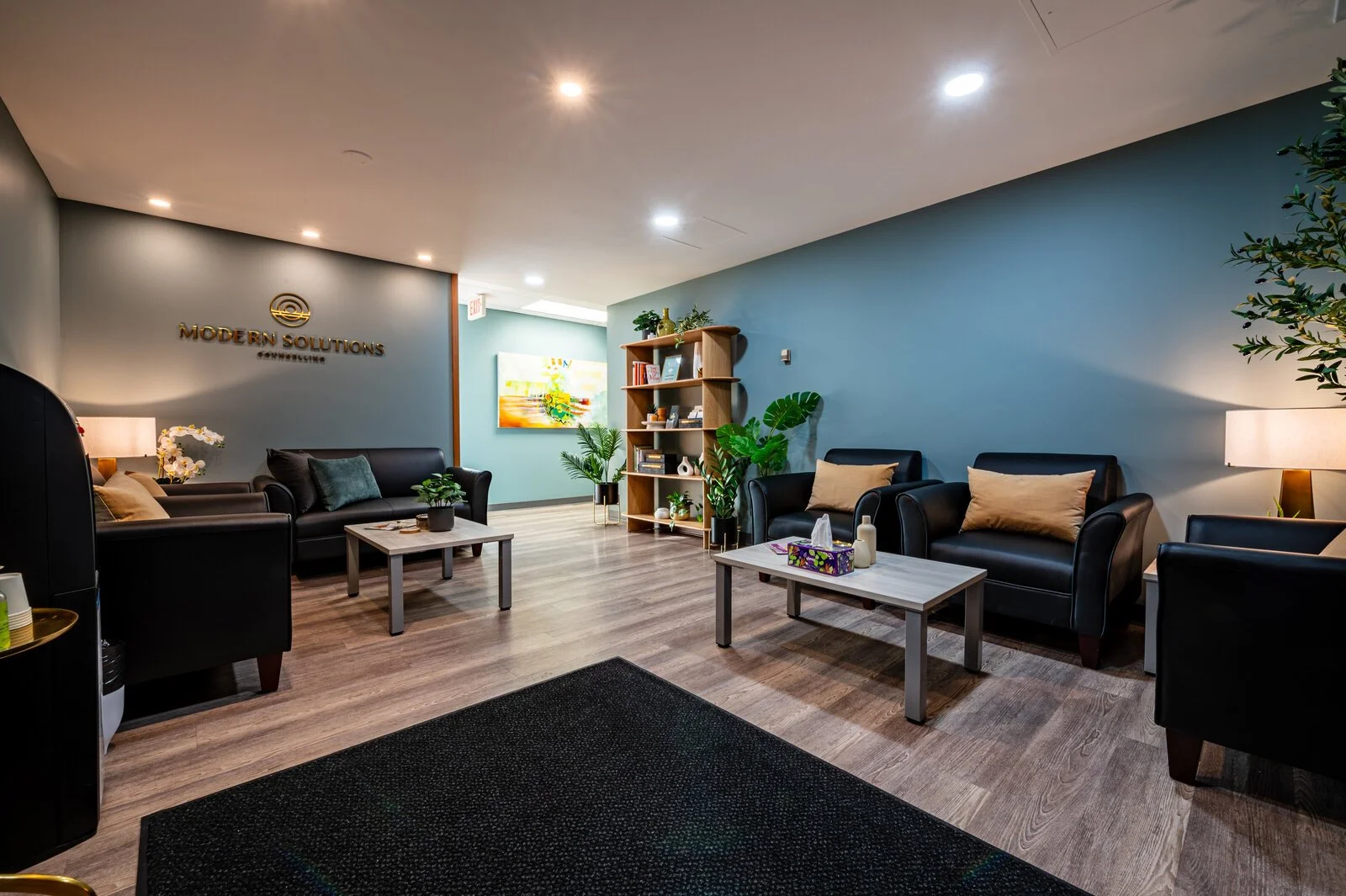 An office reception area with black leather sofas, wooden coffee tables, potted plants, and a blue accent wall with a gold Modern Solutions logo.