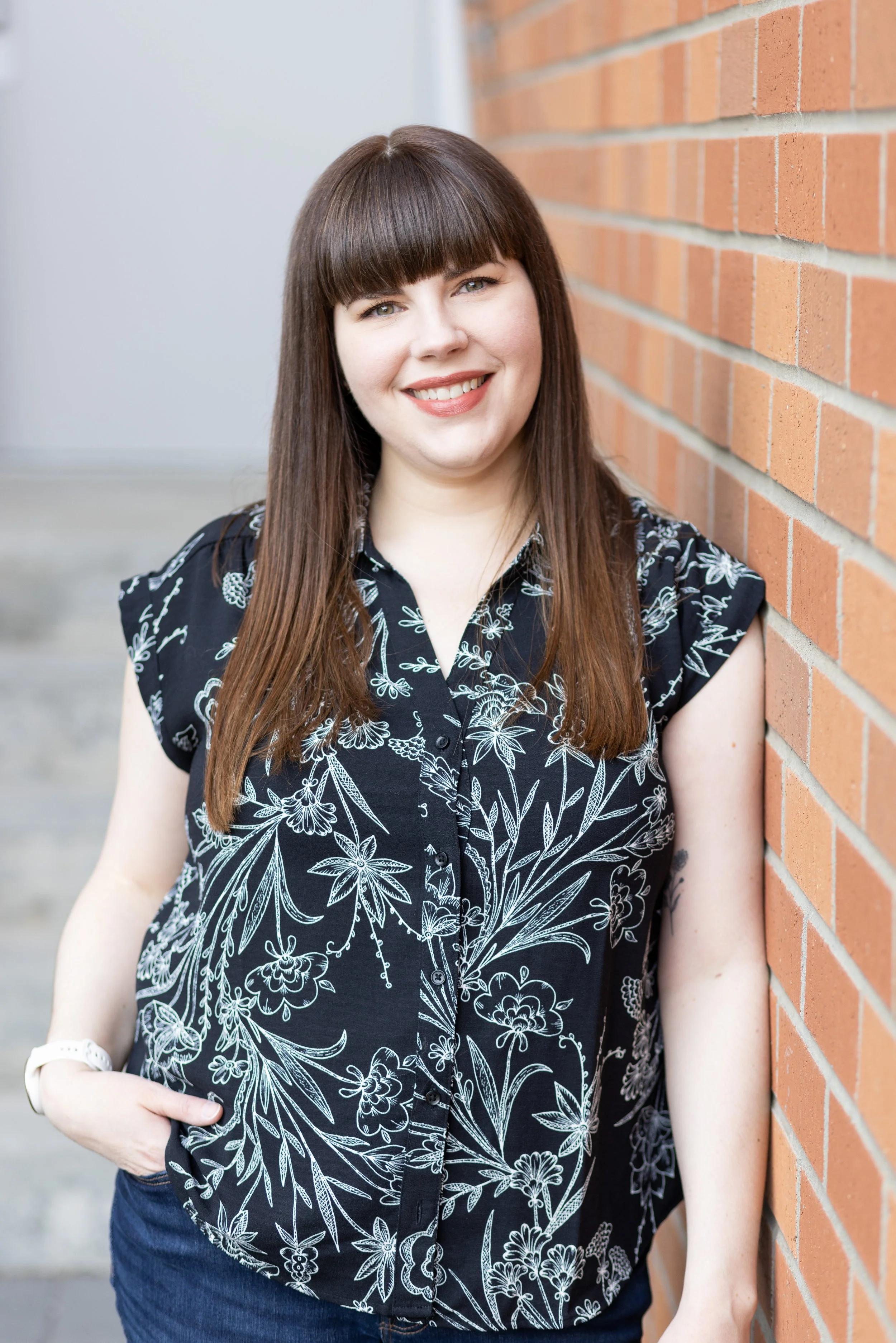 A woman with long brown hair and bangs, smiling, wearing a black shirt with white floral patterns, standing next to a brick wall.