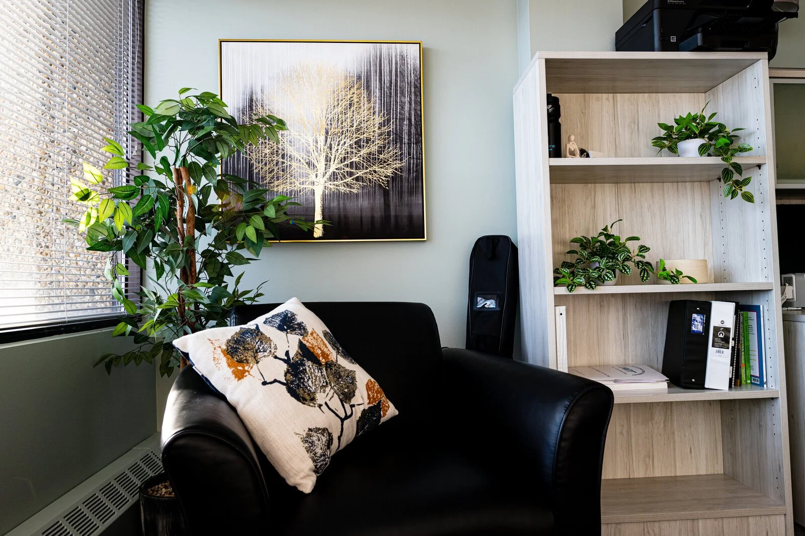 Living room corner with a window, a large potted plant, a framed art piece of a white tree, a black couch with a decorative pillow, and a wooden bookshelf with plants, books, and decorative items.