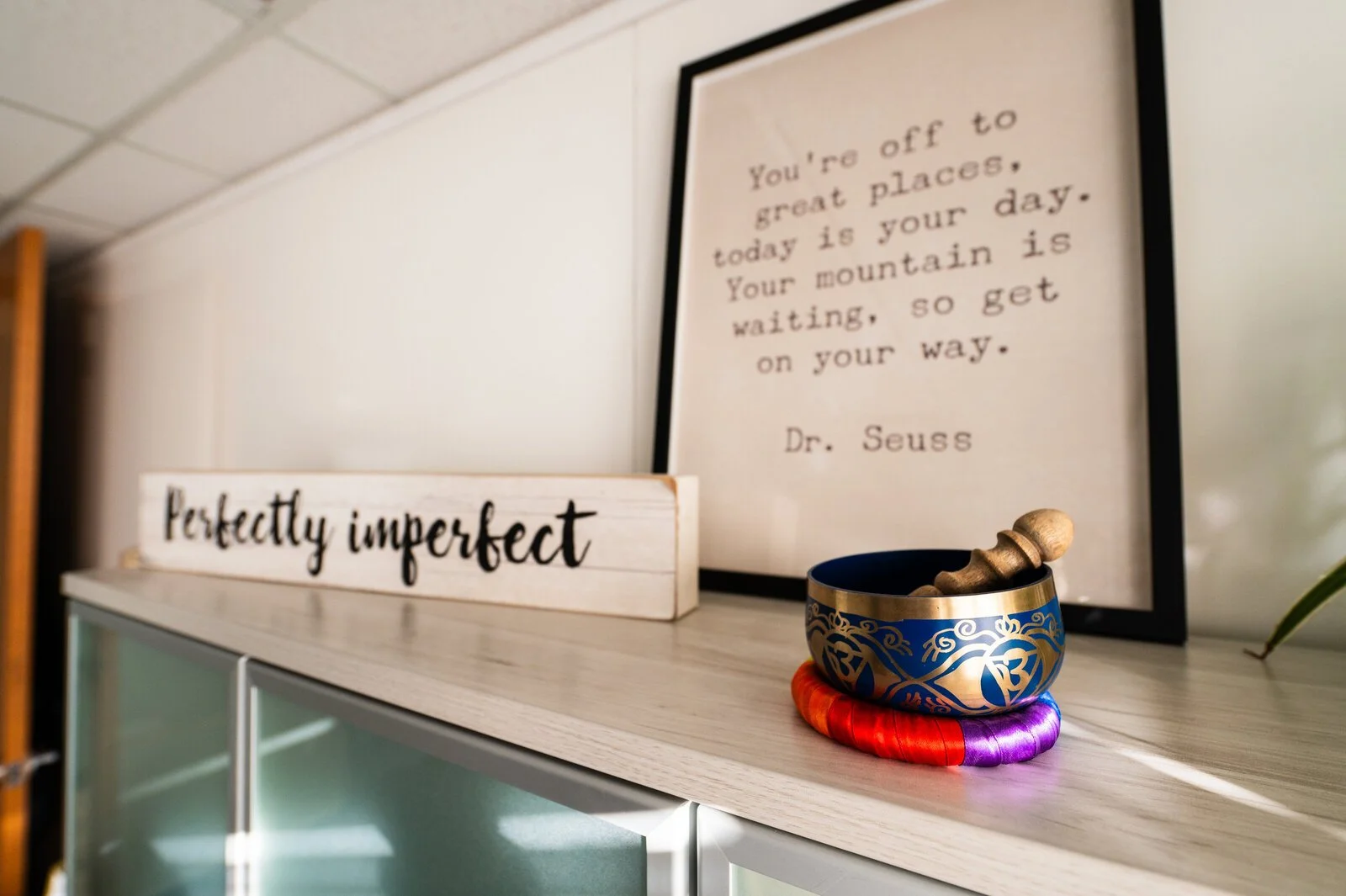 Decorative shelf with framed quote from Dr. Seuss, a colorful singing bowl with a mallet on a purple cushion, and a wooden sign reading 'perfectly imperfect'.