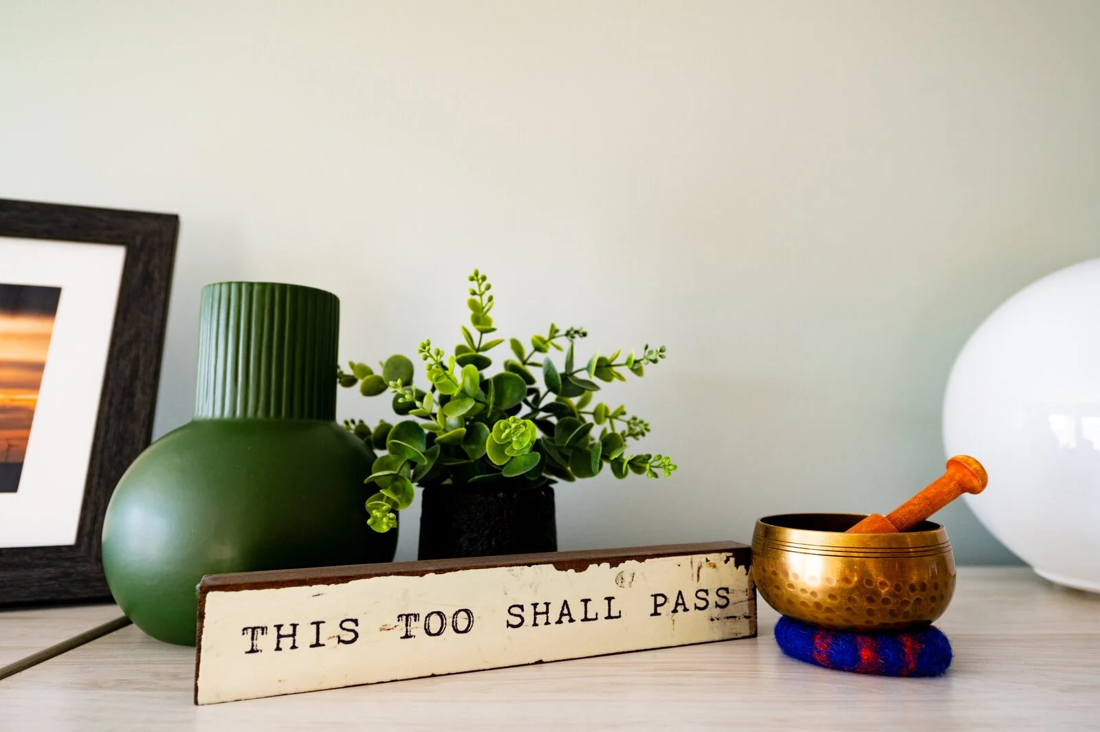 Decorative arrangement on a tabletop includes a green ribbed vase, a small potted green plant, a brass singing bowl with a wooden striker, a framed artwork, and a white spherical lamp, with a rustic sign reading "This Too Shall Pass" in the foregroun