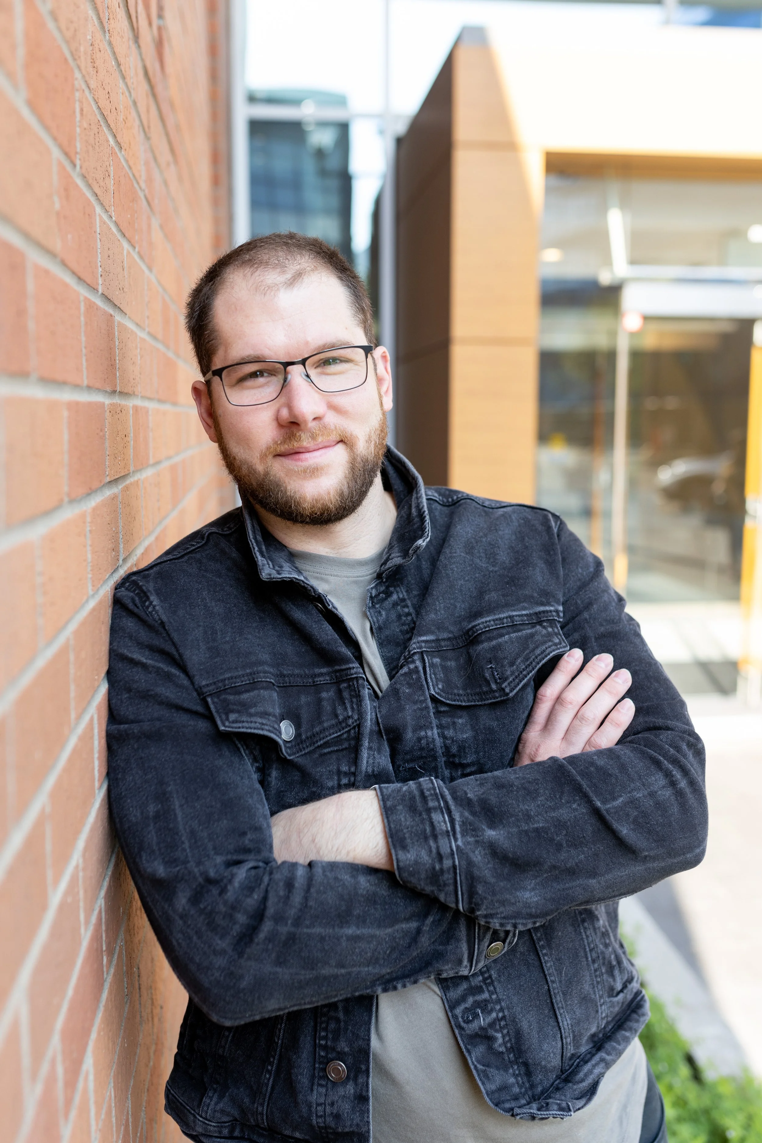 A man with glasses and a beard wearing a black denim jacket, standing with arms crossed, leaning against a brick wall outdoors.