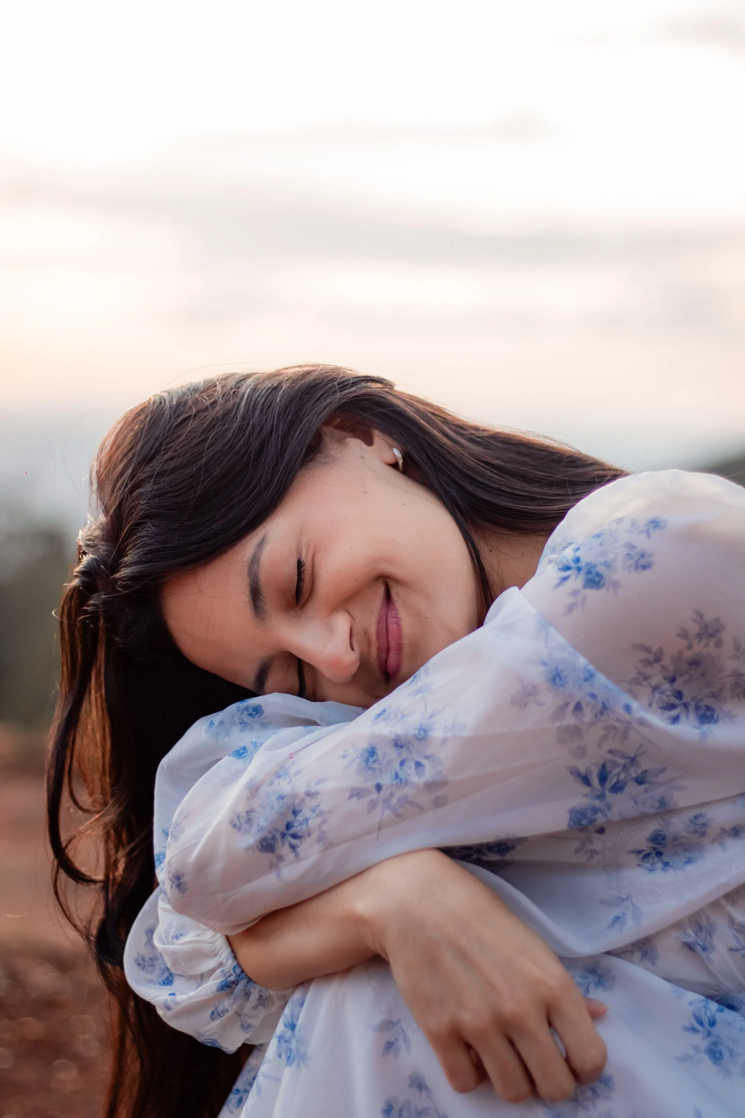 A woman with long brown hair, smiling with her eyes closed, hugging herself outdoors during sunset, wearing a white dress with blue floral patterns.