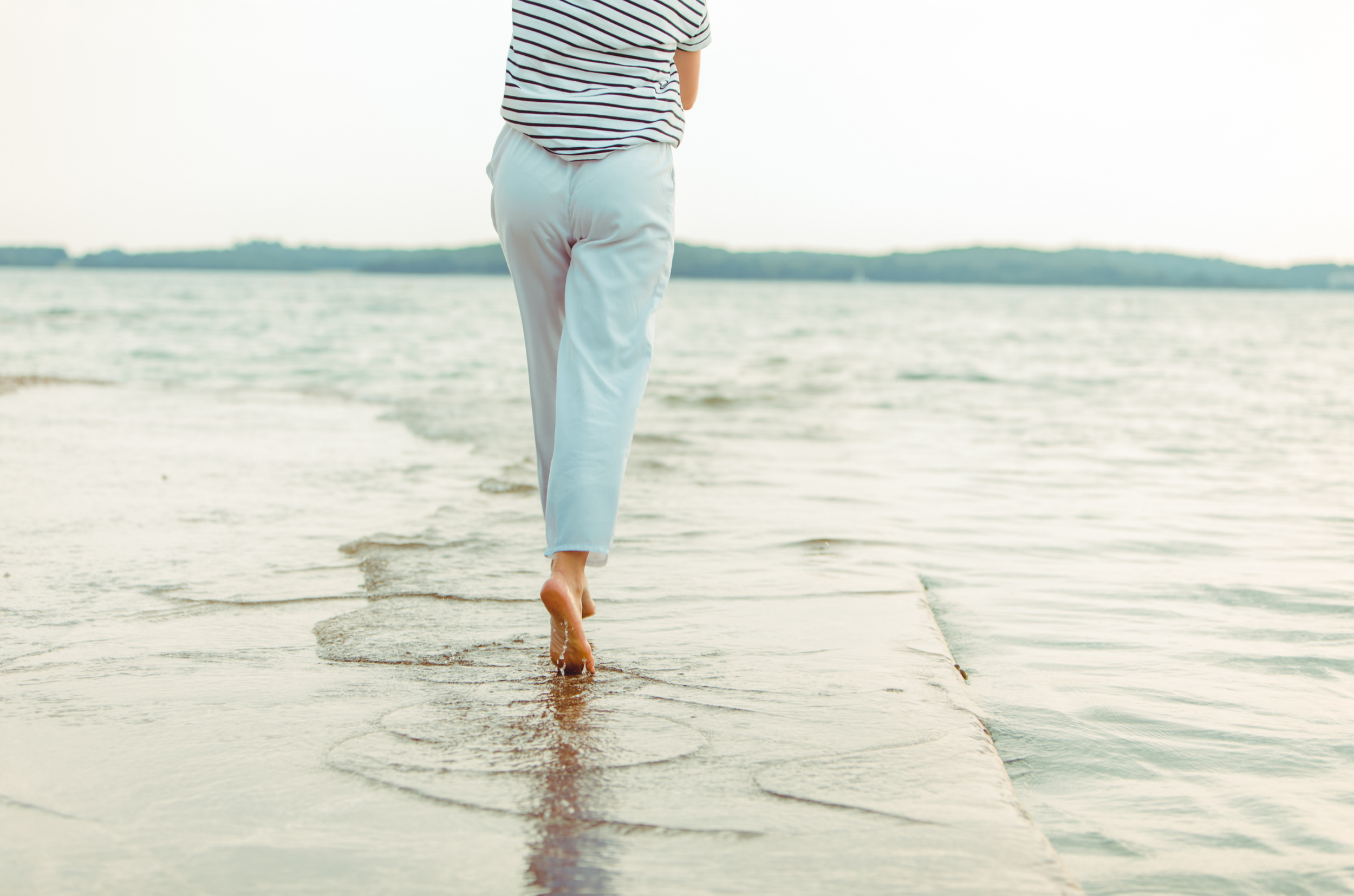A person walking barefoot on a small pier into the water at the beach, seen from behind, wearing a striped shirt and white pants.