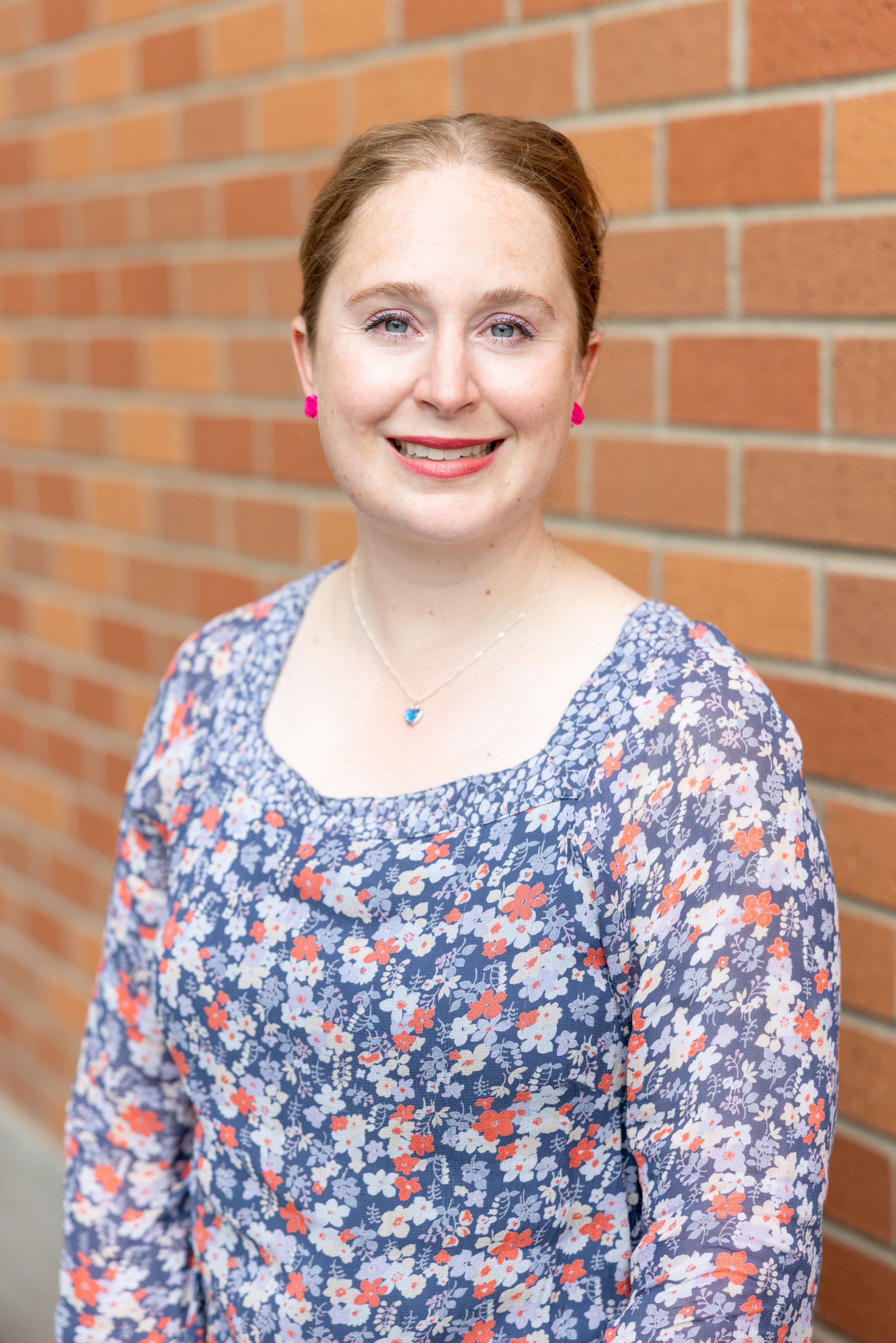 A woman with light skin, red hair, and blue eyes standing in front of a red brick wall, wearing a floral dress with pink earrings and a necklace with a heart-shaped pendant.
