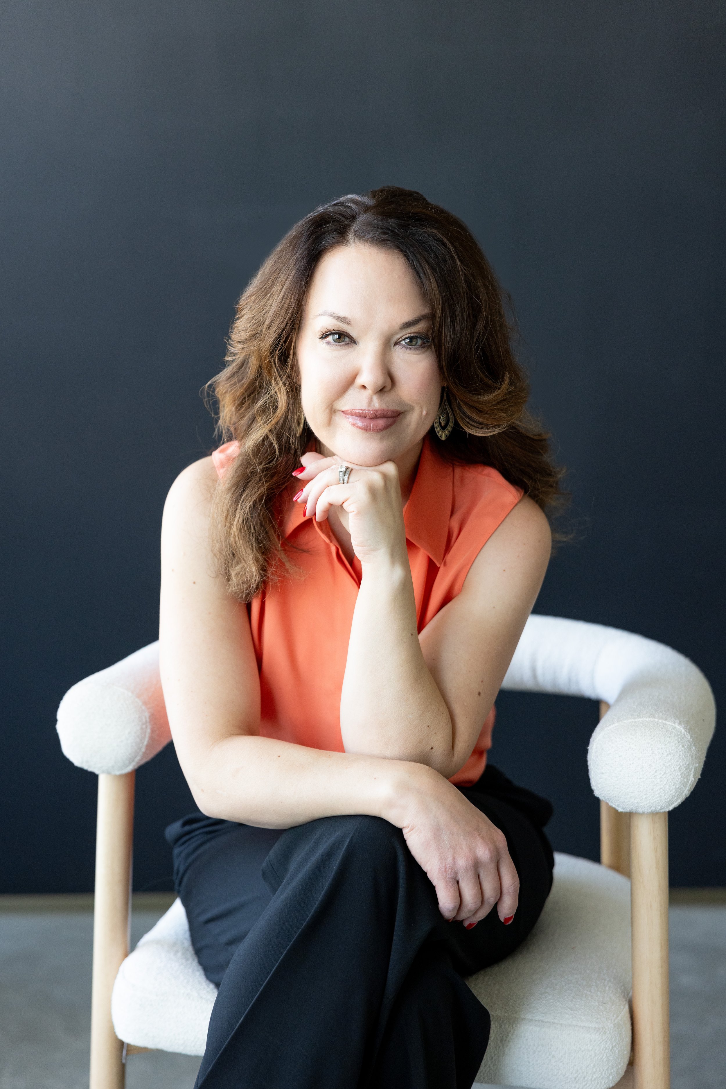 A woman with wavy brown hair, wearing an orange sleeveless top and black pants, sitting on a white upholstered chair with wooden legs, resting her chin on her hand, smiling, against a dark background.