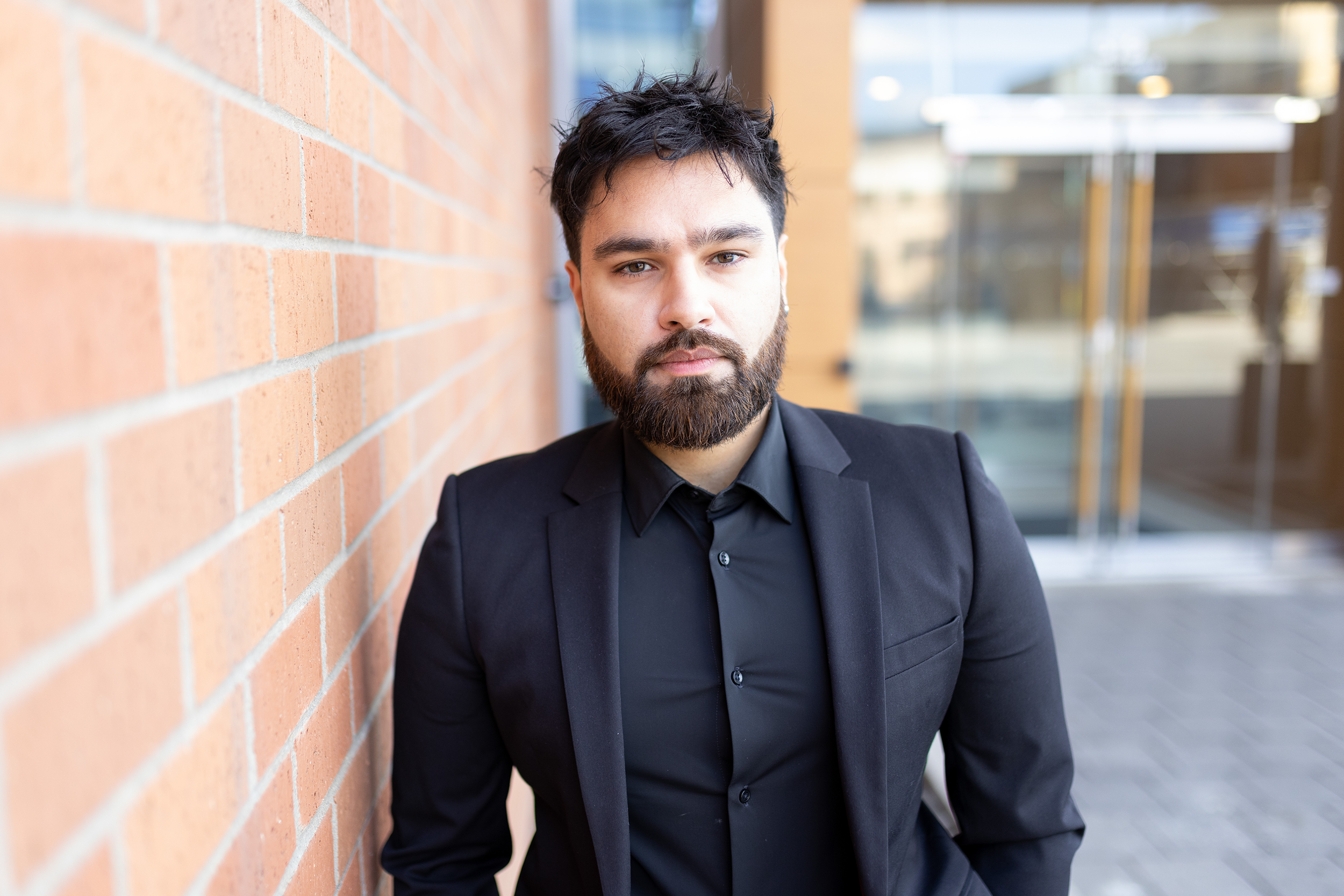 A man with dark, slightly messy hair and a beard, wearing a black suit and shirt, standing outdoors against a red brick wall with glass doors in the background.