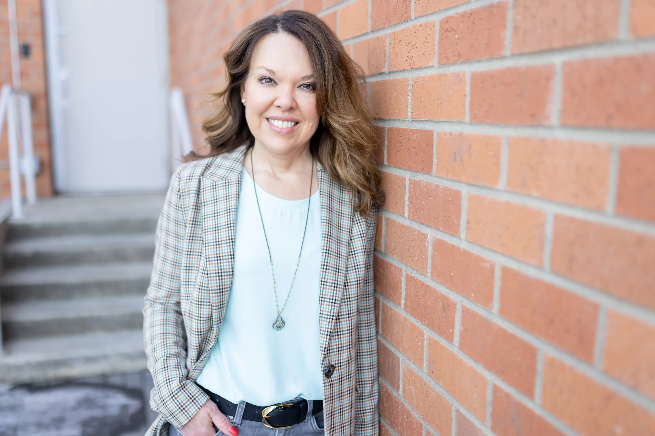 A woman with brown hair and light-colored eyes smiling while leaning against a brick wall in an urban setting.