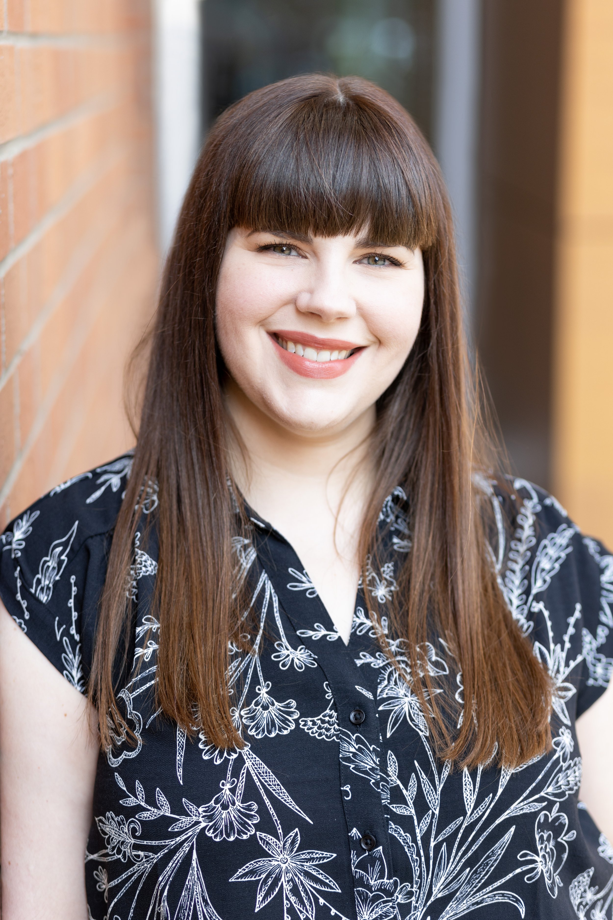 A woman with long, straight brown hair and bangs, smiling and wearing a black blouse with white floral patterns, standing outdoors against a brick wall.