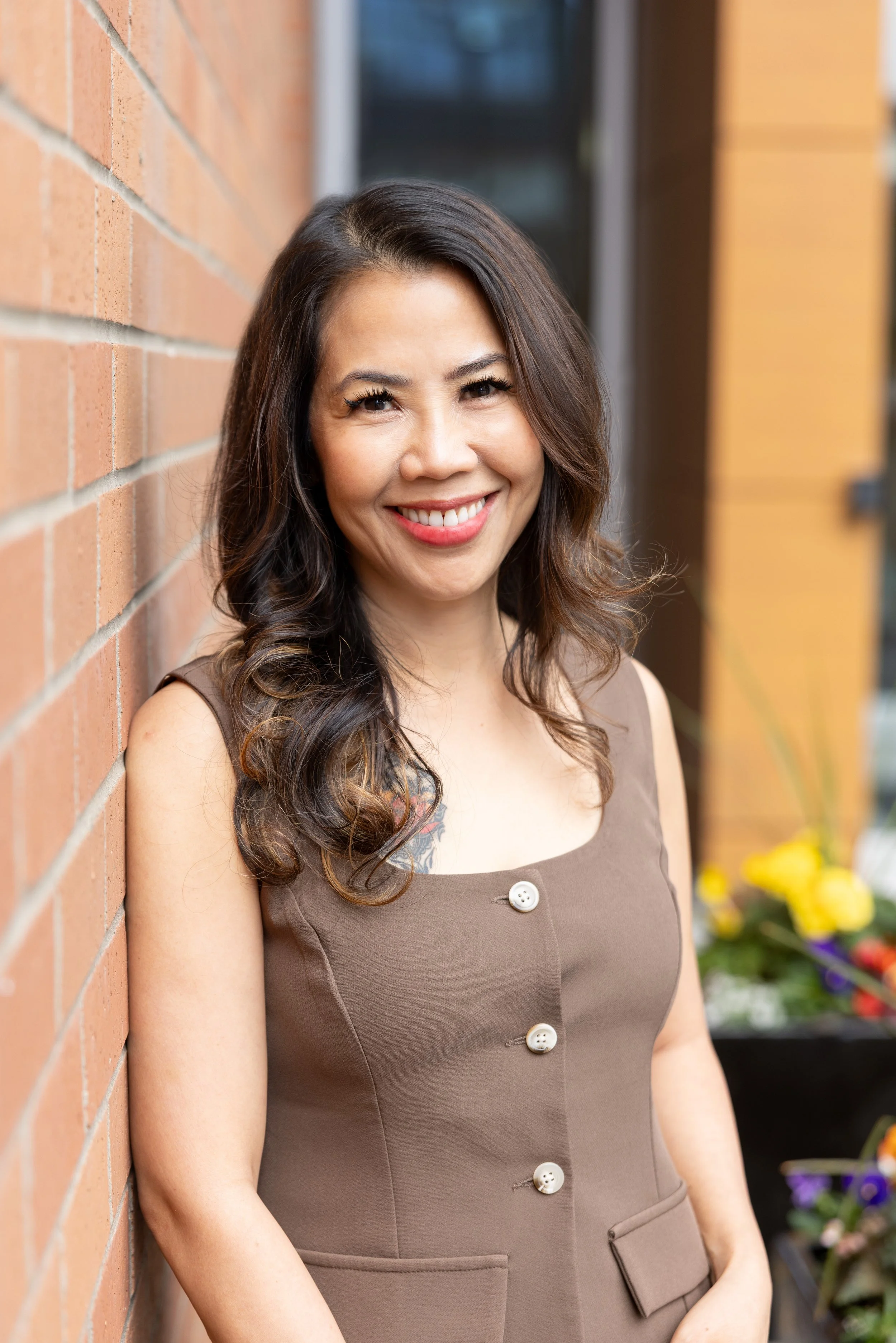 A woman with dark brown hair styled in loose waves, smiling, standing beside a brick wall. She is wearing a brown sleeveless dress with large white buttons and front pockets. In the background, there are colorful flowers.