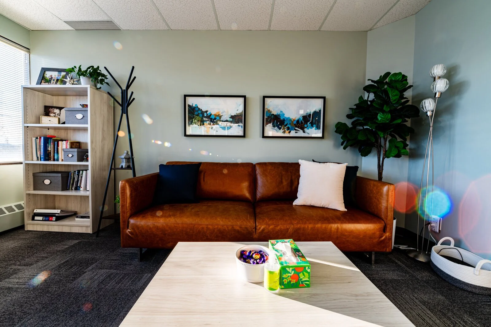 Living room with a brown leather sofa, two colorful abstract paintings on the wall, a white coffee table with decorative items, a tall houseplant, a white bookshelf filled with books and storage boxes, and a multi-globe floor lamp. Sunlight coming th