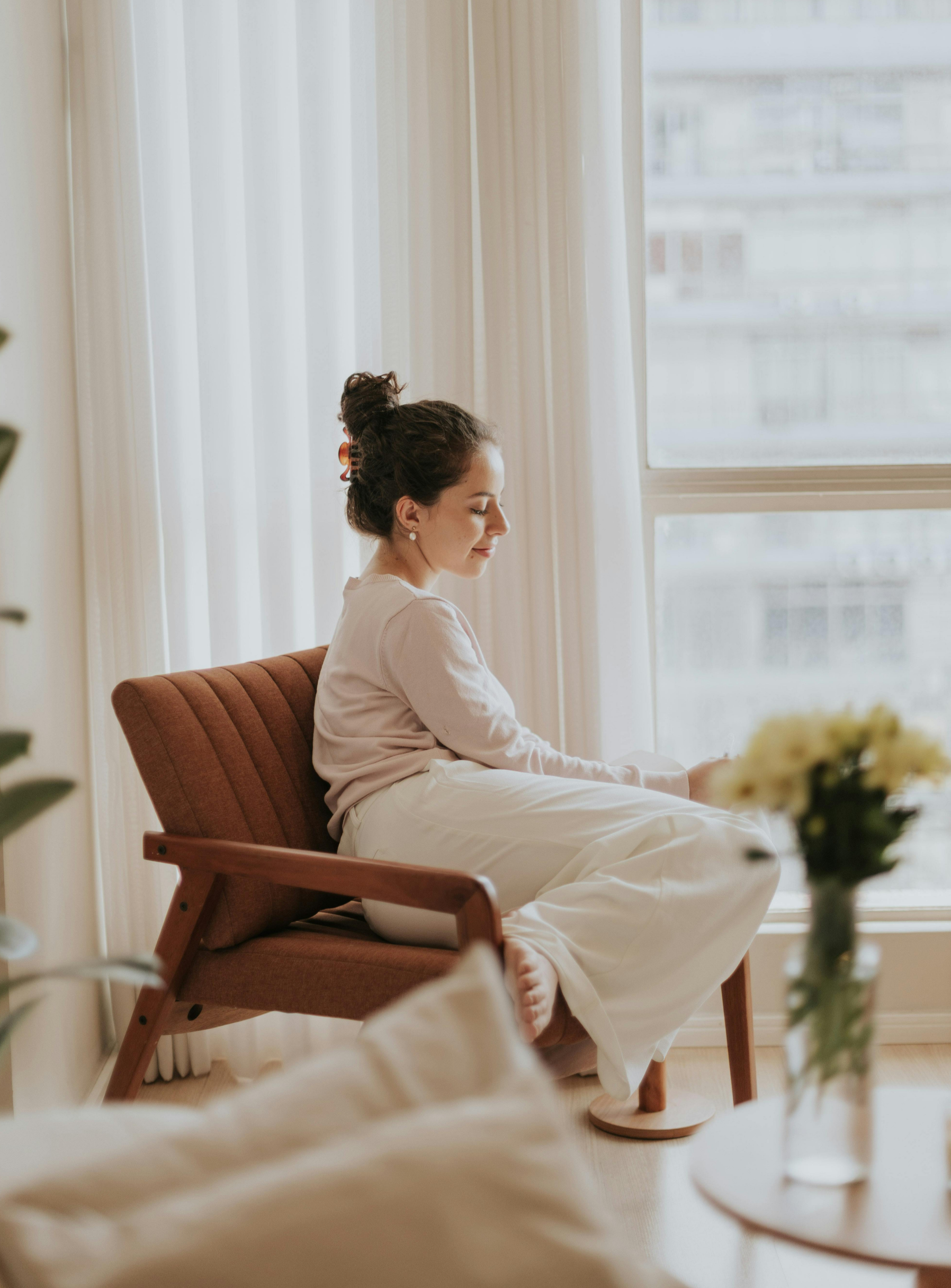 A woman sitting calmly on a mid-century modern armchair indoors near a large window with white curtains, dressed in light-colored clothing, with her eyes closed and a peaceful expression.