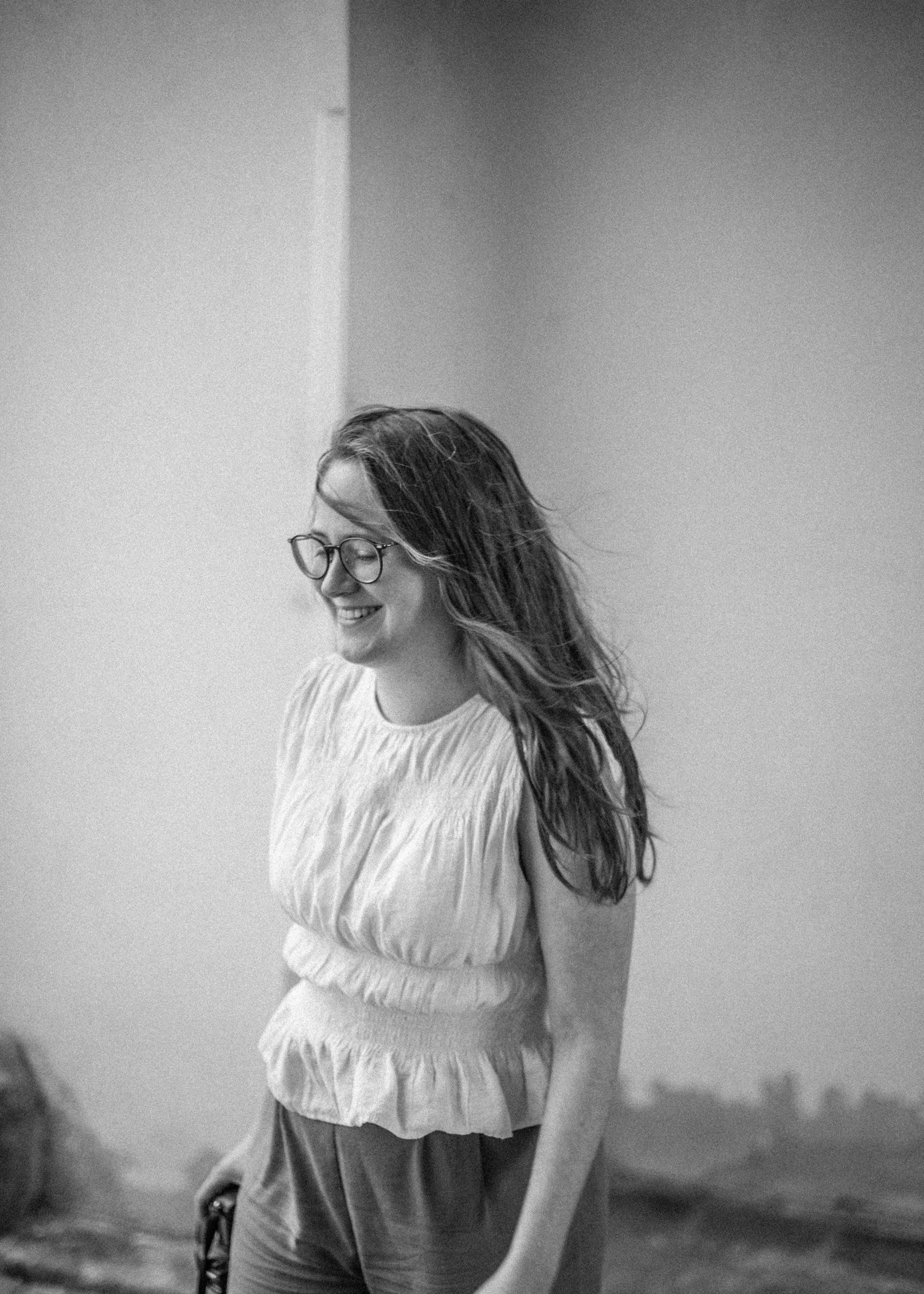 Ein lachendes Mädchen mit Brille, das eine schlichte, kurze, geraffte Bluse trägt, in einer Ecke eines Raumes mit hellgelbem Boden. Das Bild ist in Schwarz-Weiß gehalten.