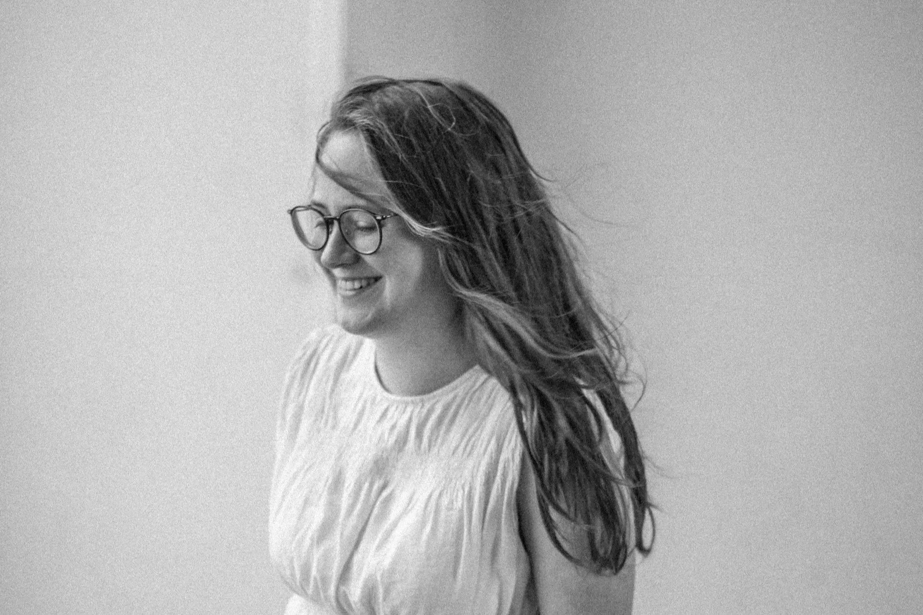 Eine lächelnde junge Frau mit lockigem Haar und Brille, die nach rechts blickt, vor einer hellen, schlichten Wand.