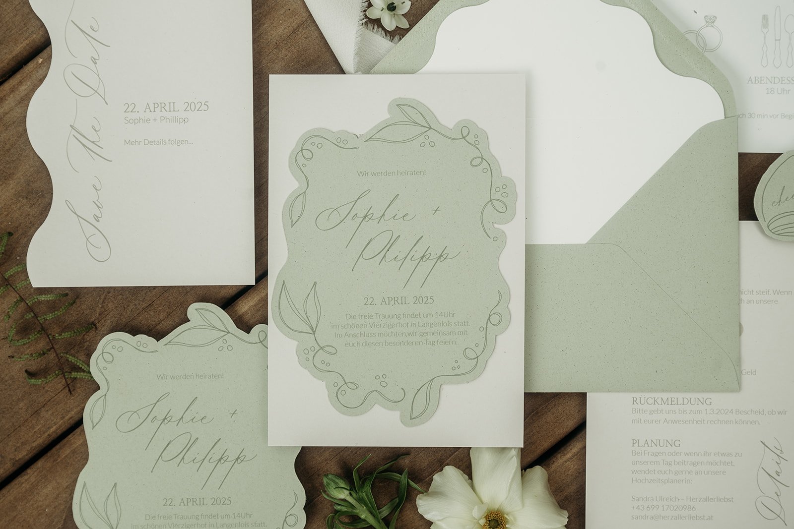 Hochzeitseinladungen mit grünen und weißen Akzenten, Blätter- und Design-Elementen, auf einem Holztisch mit Blumen. Ein Einladungsschild zeigt die Namen Sophie und Philipp, die Hochzeit am 22. April 2025. Weitere Einladungen und Briefumschläge sind sichtbar.