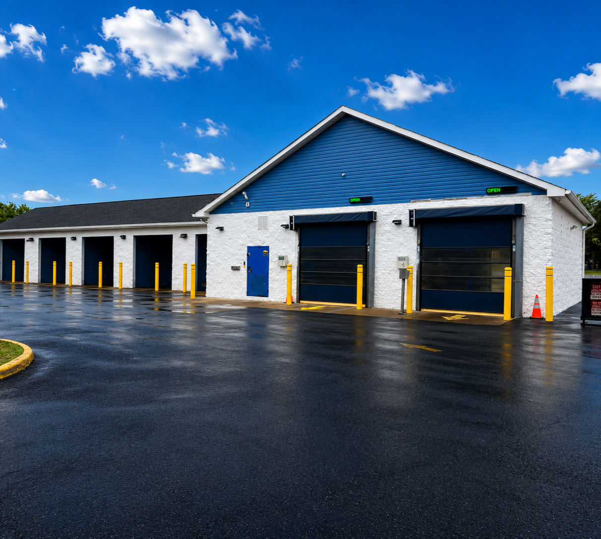 A clean, empty parking garage with four large blue garage doors, yellow safety bollards, and a blue and white building exterior under a bright blue sky with scattered clouds.