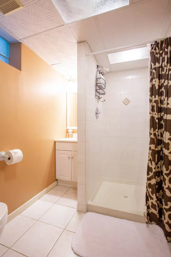 Bathroom with a shower stall, brown and white patterned curtain, white tiles, yellow wall, vanity with mirror and lotion, toilet paper holder with roll, white tile floor, and skylight.