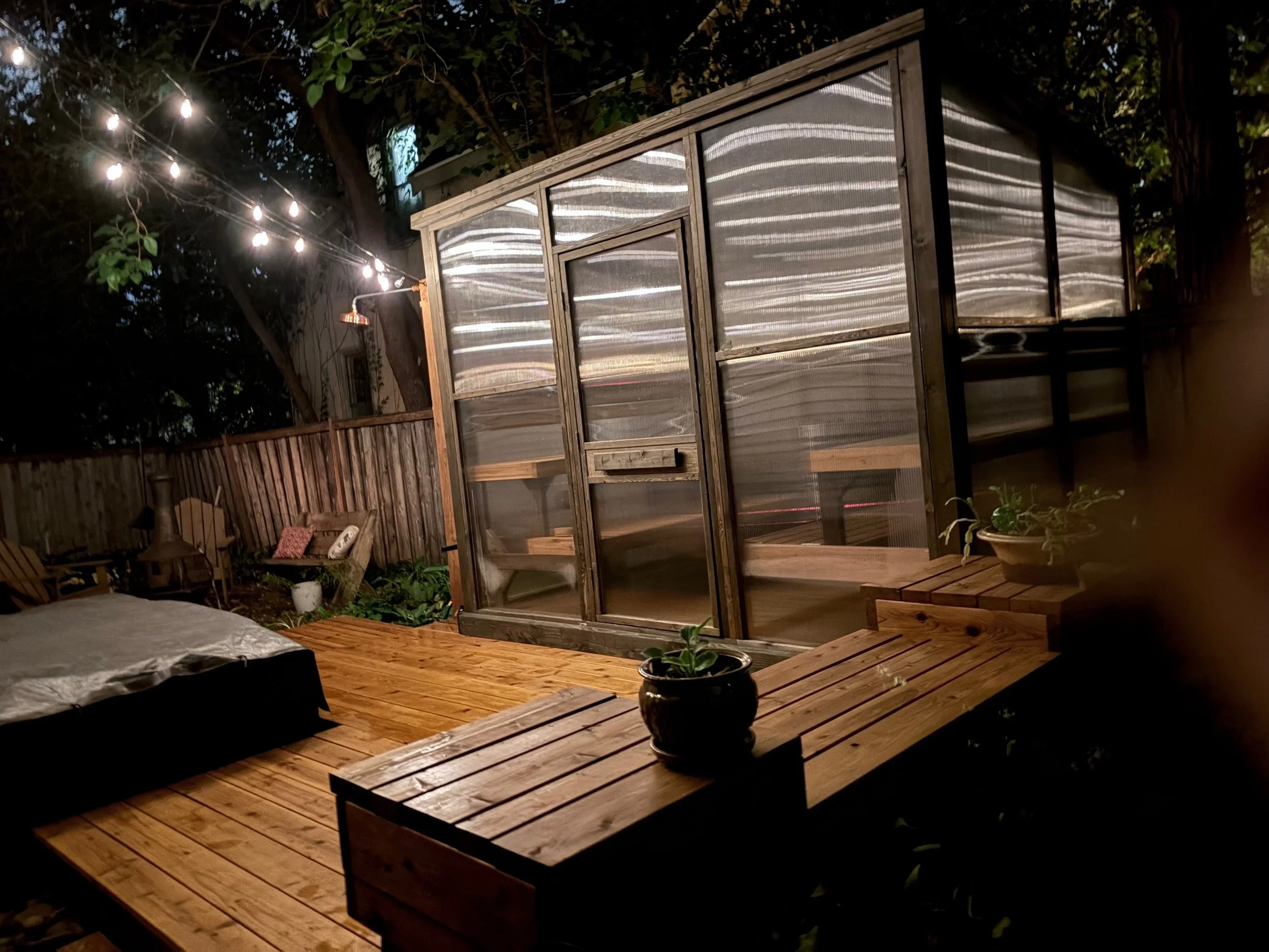 A backyard deck at night with string lights hanging from trees, a small greenhouse, potted plants, and wooden furniture.