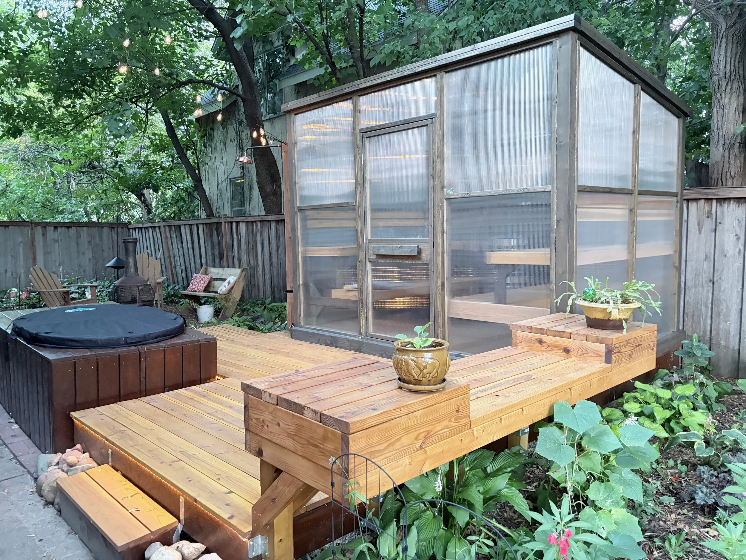 Wooden deck with potted plants, a black fire pit, and outdoor furniture, enclosed by a wooden fence with trees and string lights overhead.