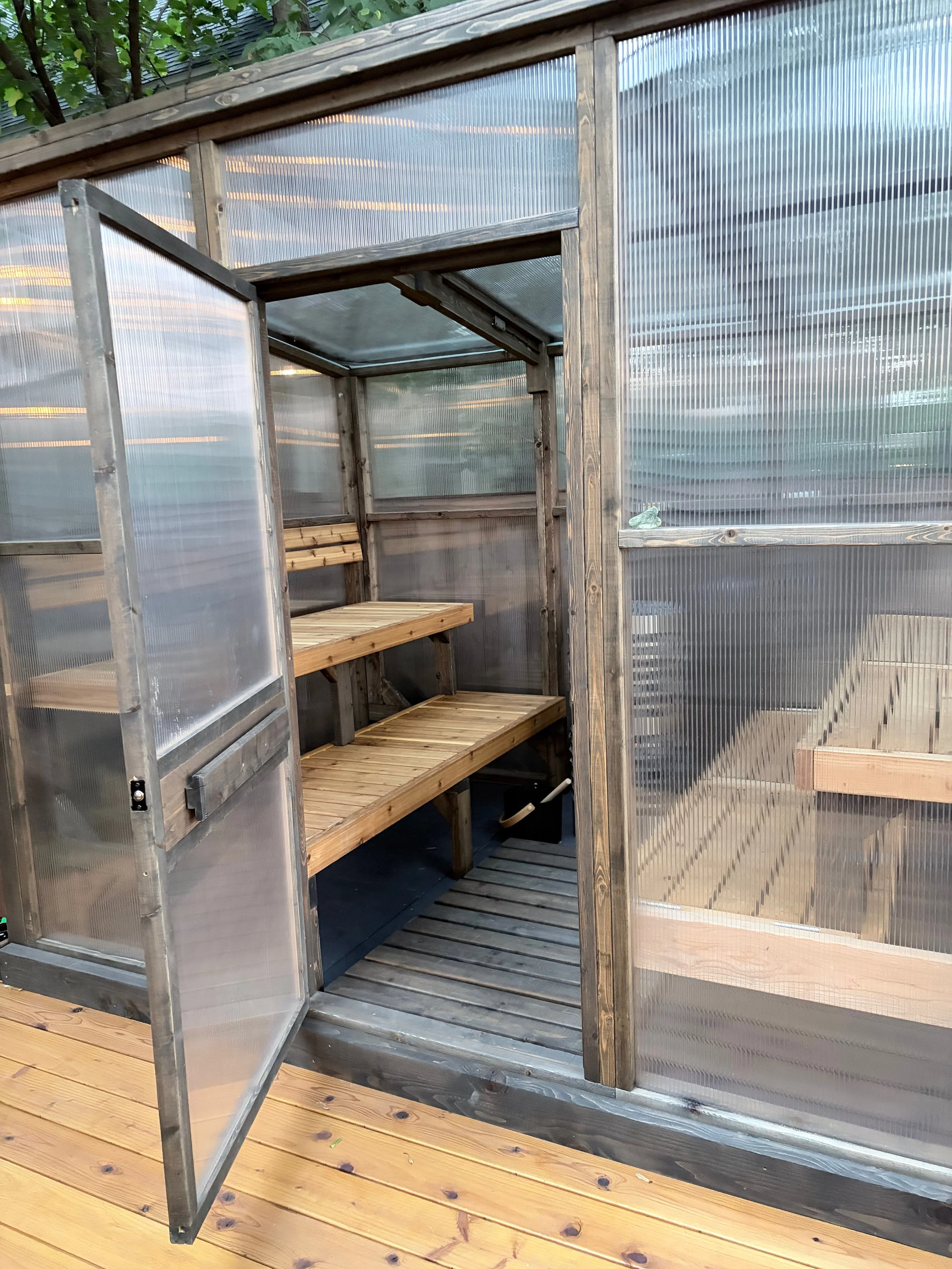 A small greenhouse with wooden shelves and a semi-transparent plastic cover, sits on a wooden deck outdoors.