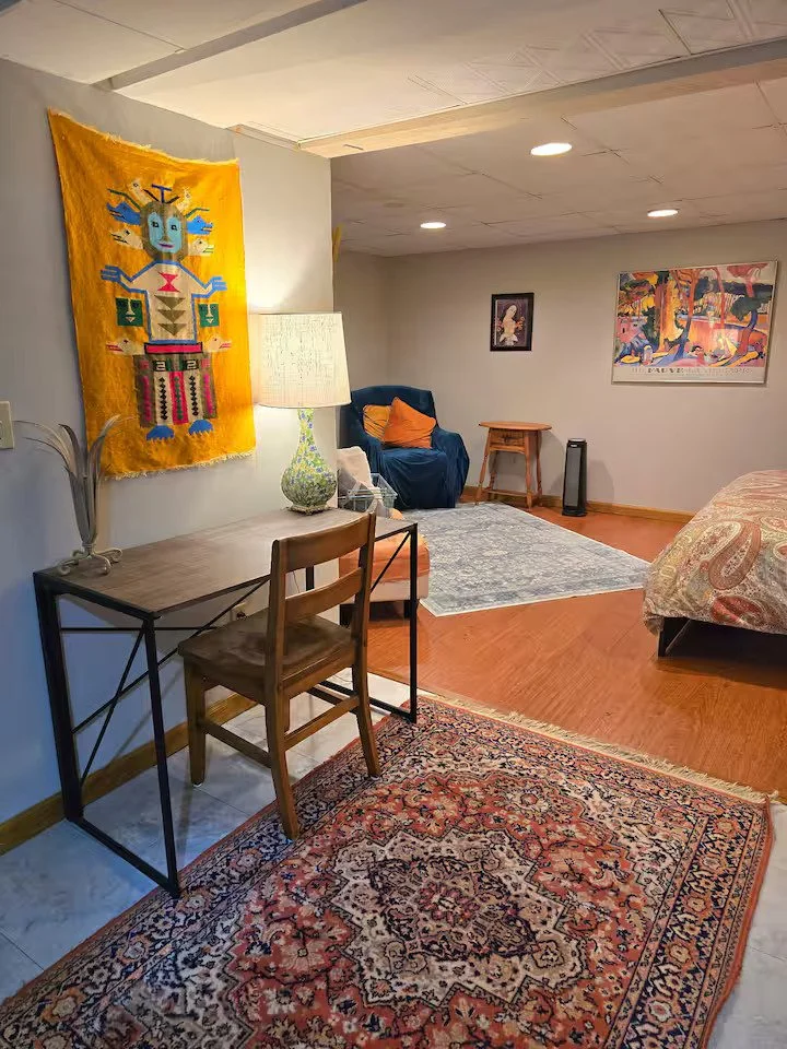 Living room with a wooden desk and chair, colorful tribal tapestry on the wall, table lamp, armchair with orange pillow, artwork, area rug, bed partially visible.