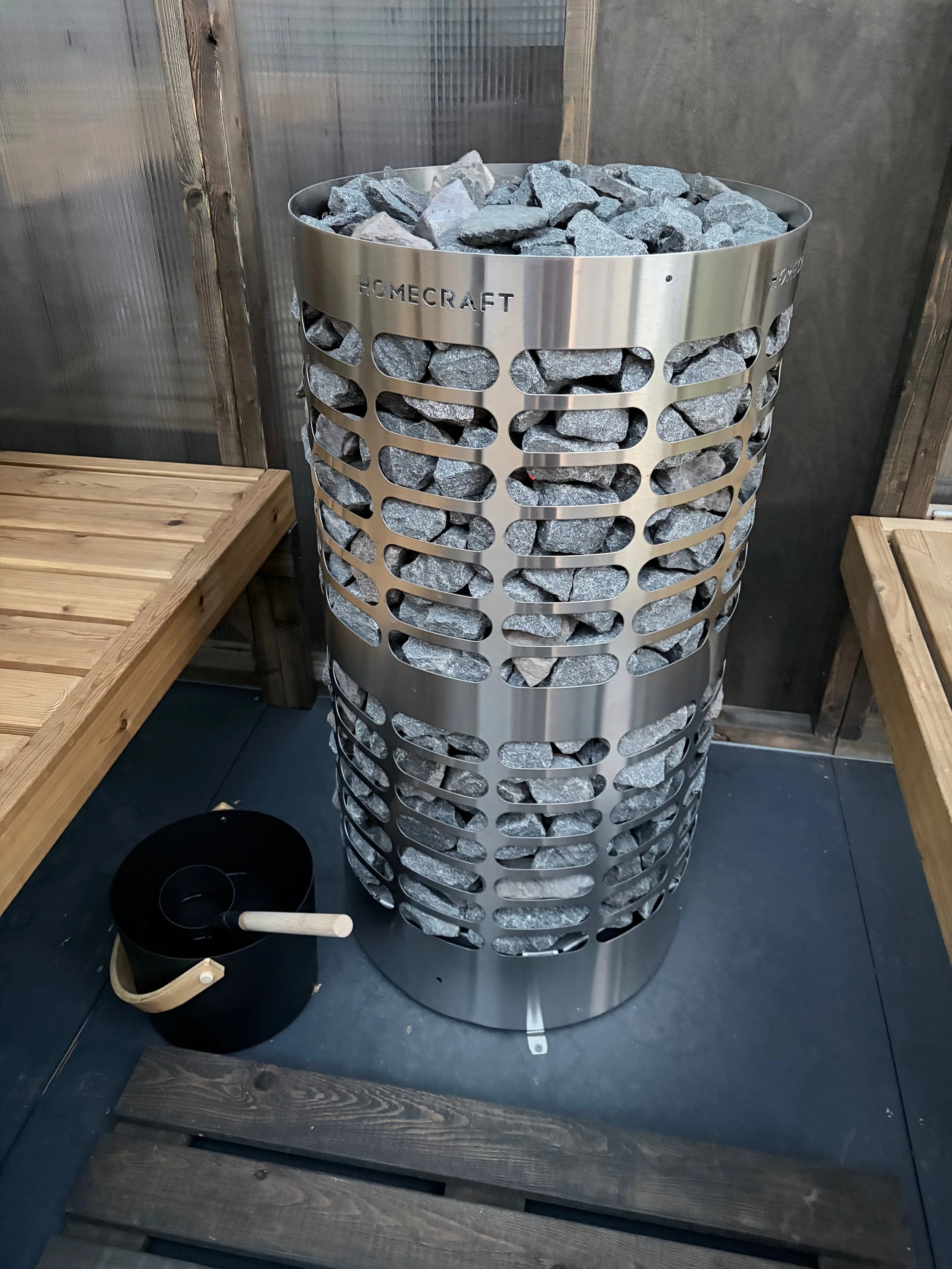 A metallic sauna heater filled with gray stones, placed on a black floor with wooden benches nearby.