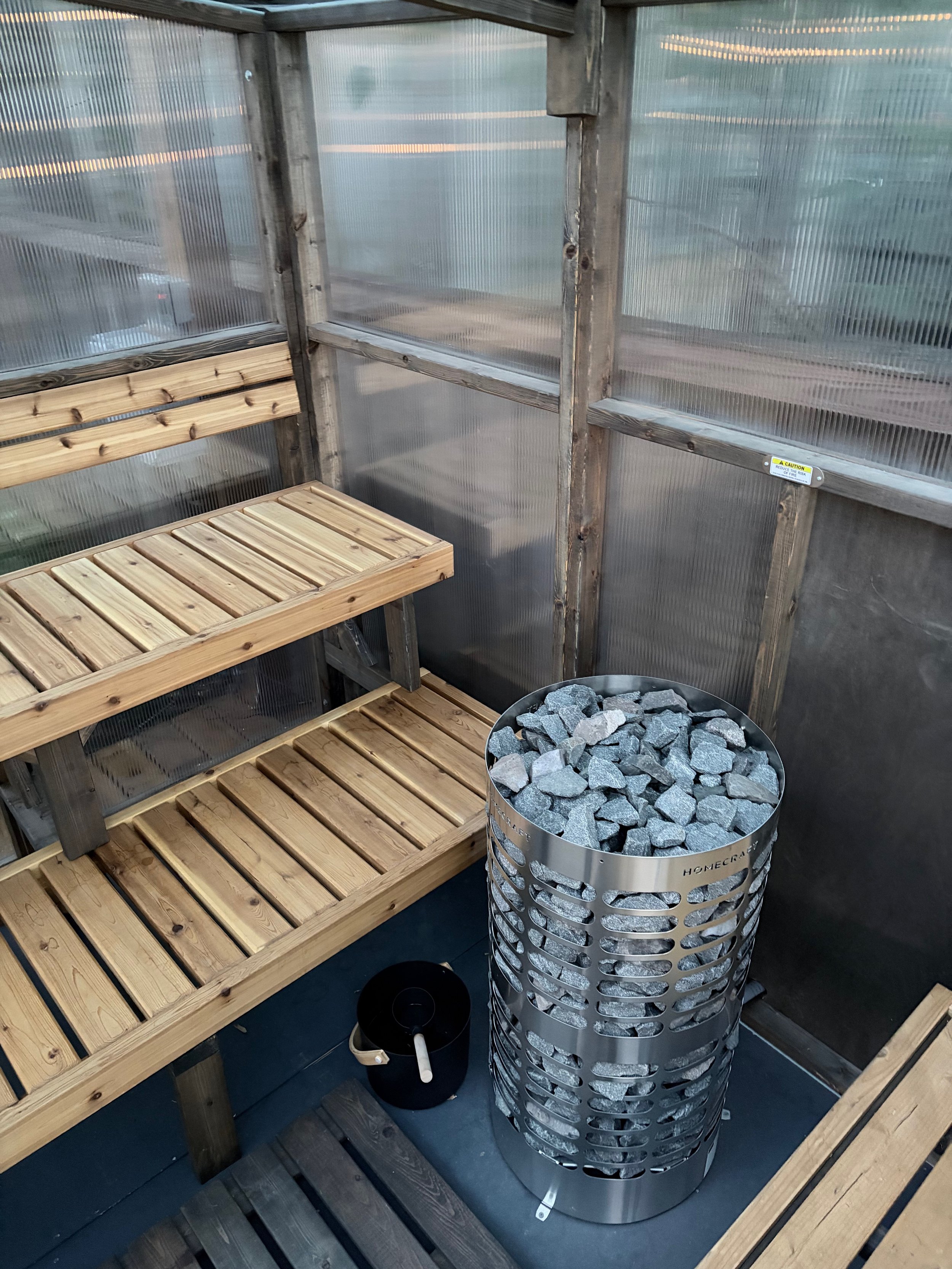 A cedar sauna with translucent walls with wooden benches lining the interior walls, an electric sauna heater filled with rocks, and a small black bucket on the floor.