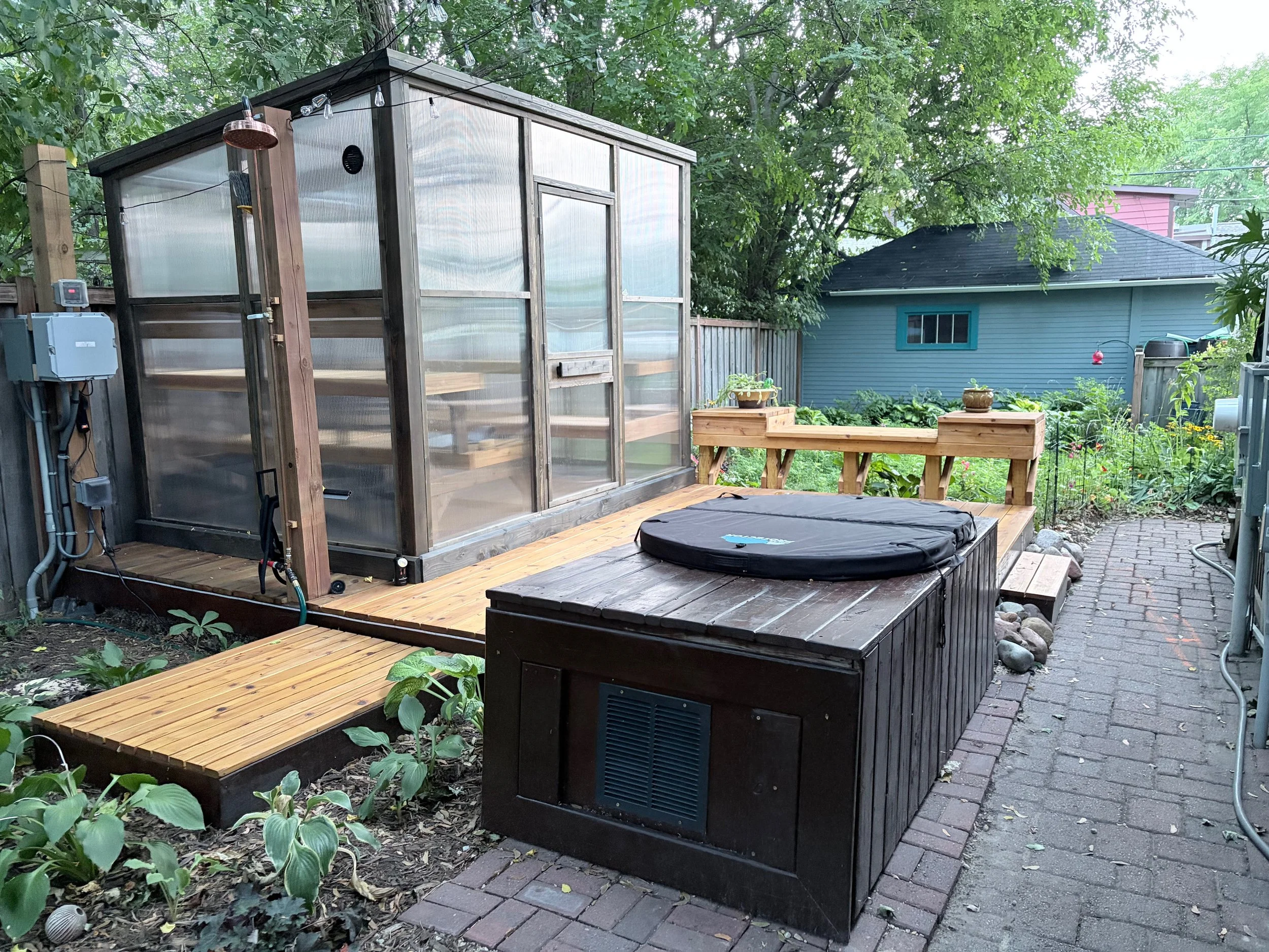 Backyard with a cedar sauna with translucent walls sitting on a wooden deck next to a cold plunge tub, surrounded by a garden with plants, a pathway, and trees.