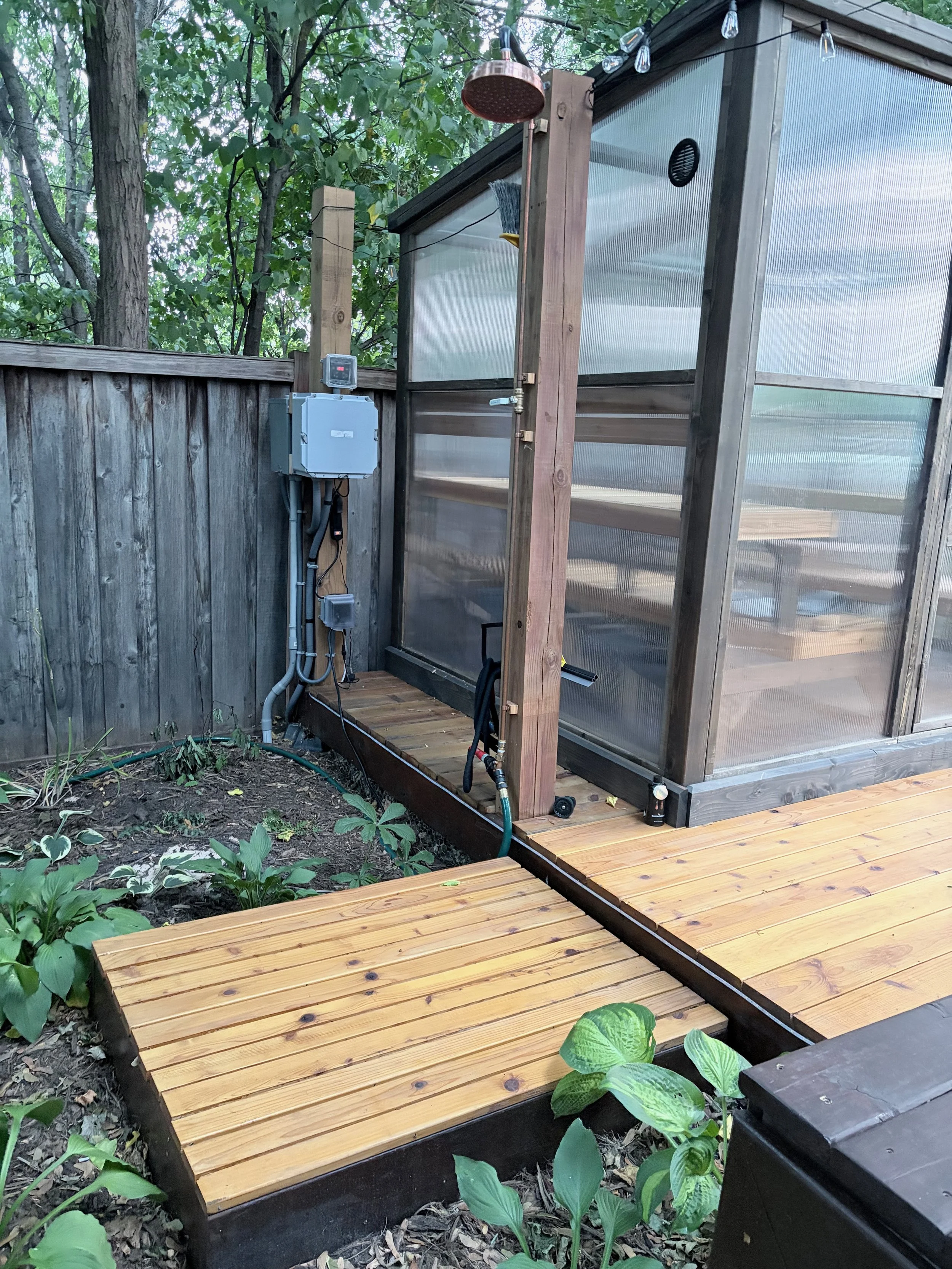 A backyard scene showing a wooden deck, a small garden, a cedar sauna with translucent walls and an outdoor shower with a copper rainfall shower head and some string lights hanging above. The surrounding area has trees and green plants.