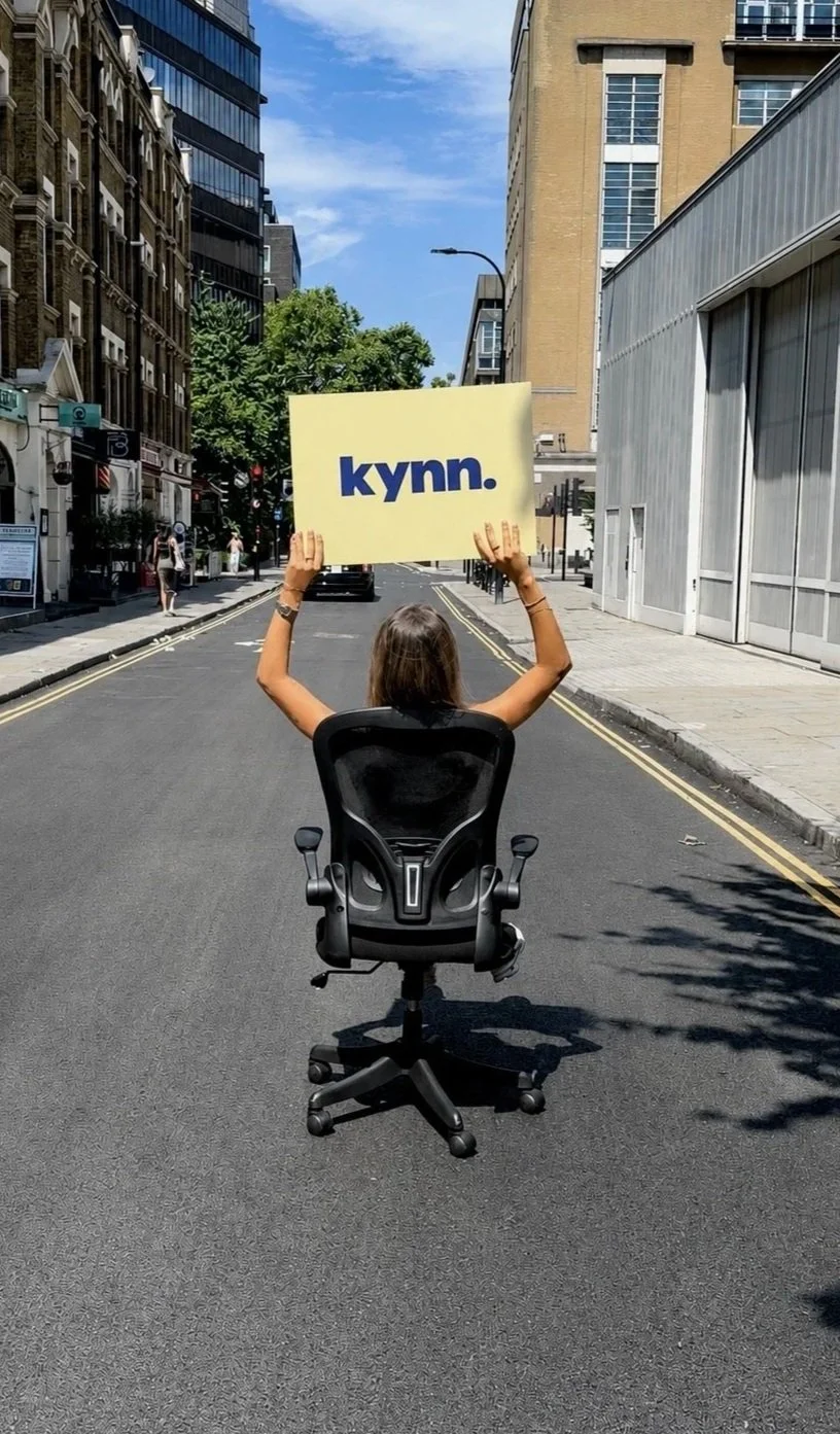 Person sitting on office chair in the middle of a city street, holding a sign that says 'kynn.'.