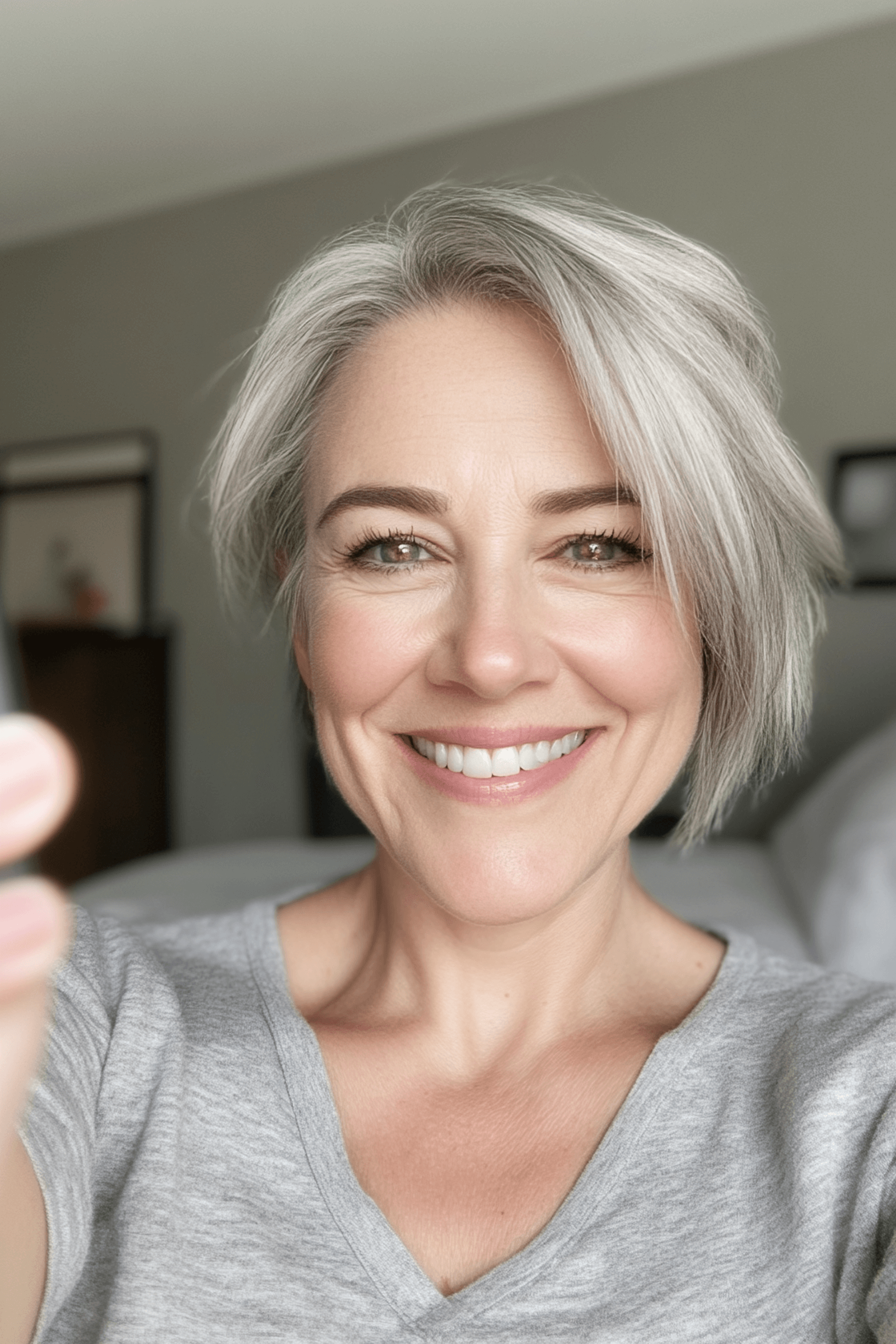 Une femme souriante à cheveux gris prisant un selfie dans une pièce lumineuse.