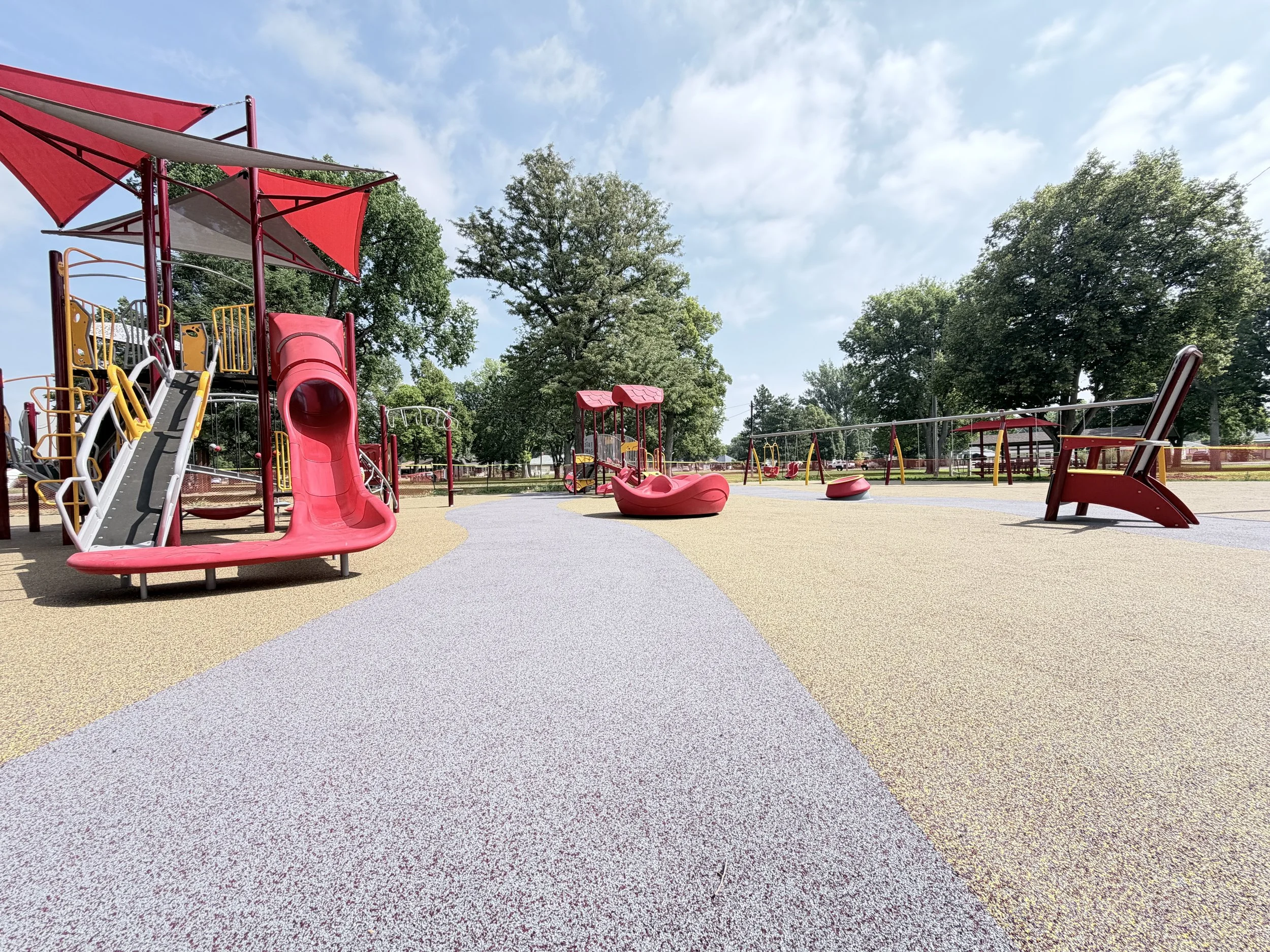 An empty playground with red slides, swings, a sandbox, and benches, surrounded by green trees and a blue sky with some clouds.