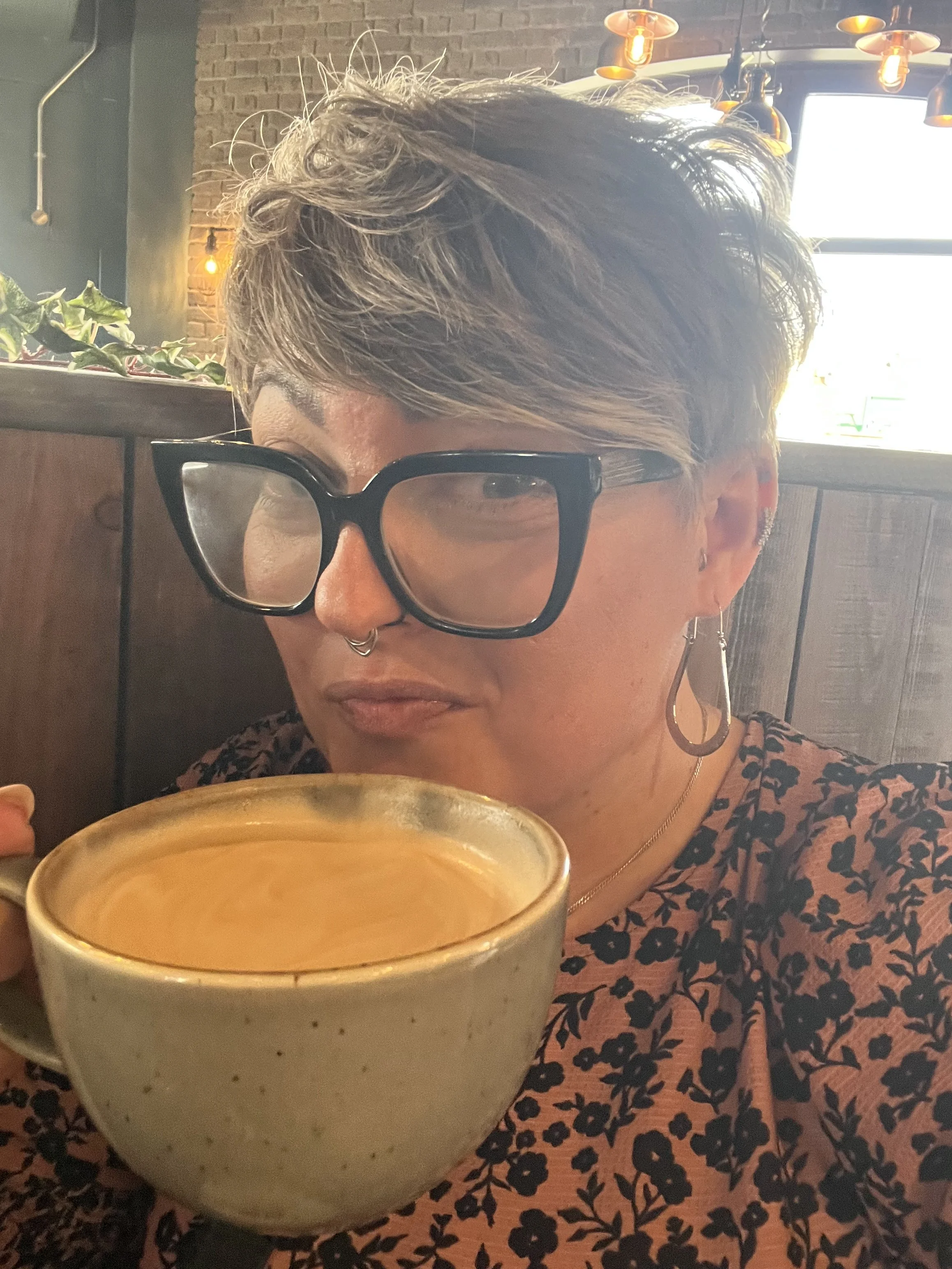Ria Jackson with short, silver hair and glasses sitting in a cafe, holding a cup of coffee. The cafe has brick walls and hanging orange lamps.
