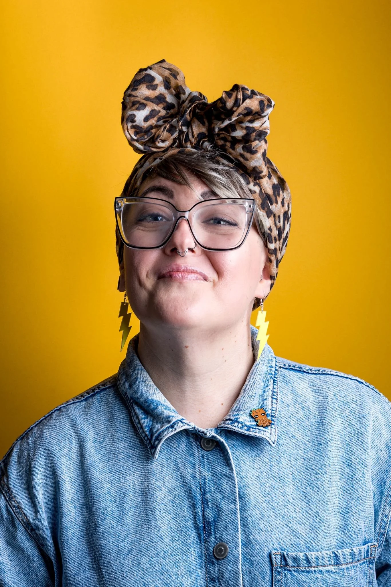 Ria Jackson with short hair wearing glasses, a leopard print headscarf tied into a large bow on top, and a denim dress with a Groot pin, standing against a yellow background. Photograpy by Joanna Wood