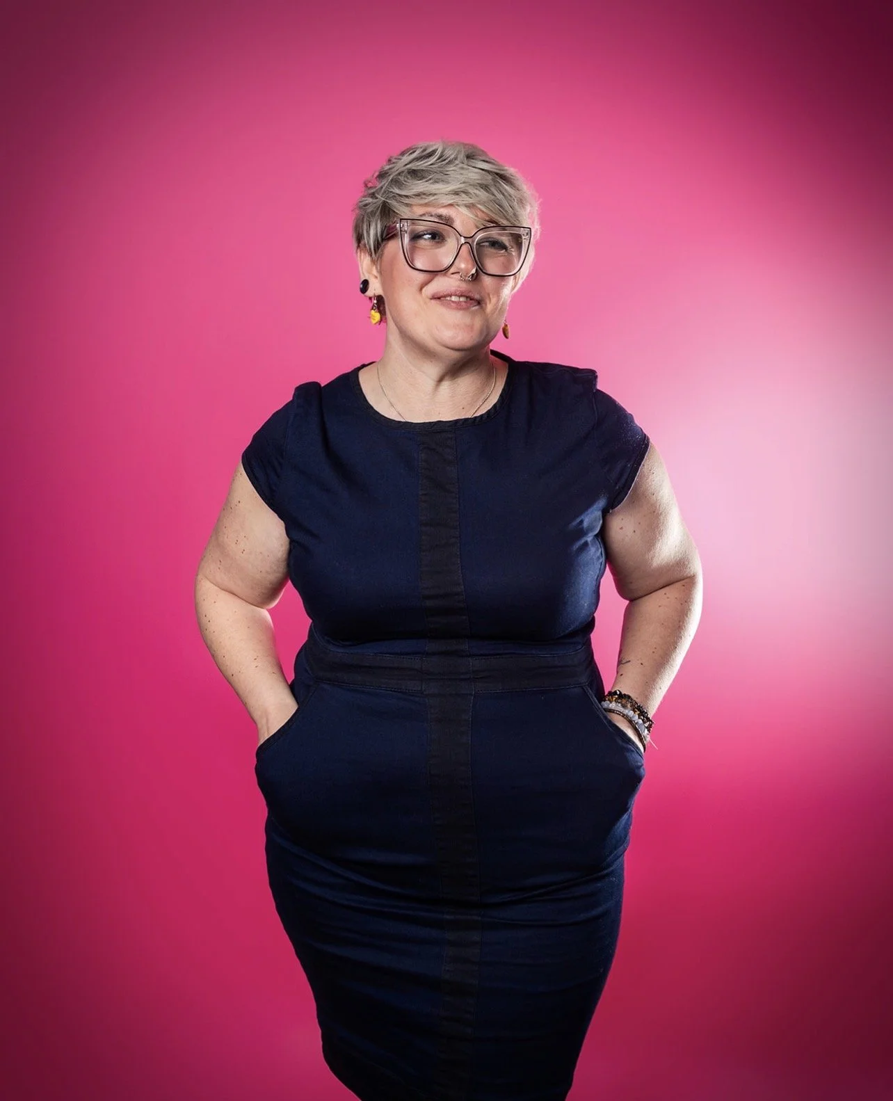 Ria Jackson with short silver hair and glasses posing against a pink gradient background. She is wearing a dark blue dress with her hands in her pockets, smiling softly. Photography By Haydn Brown