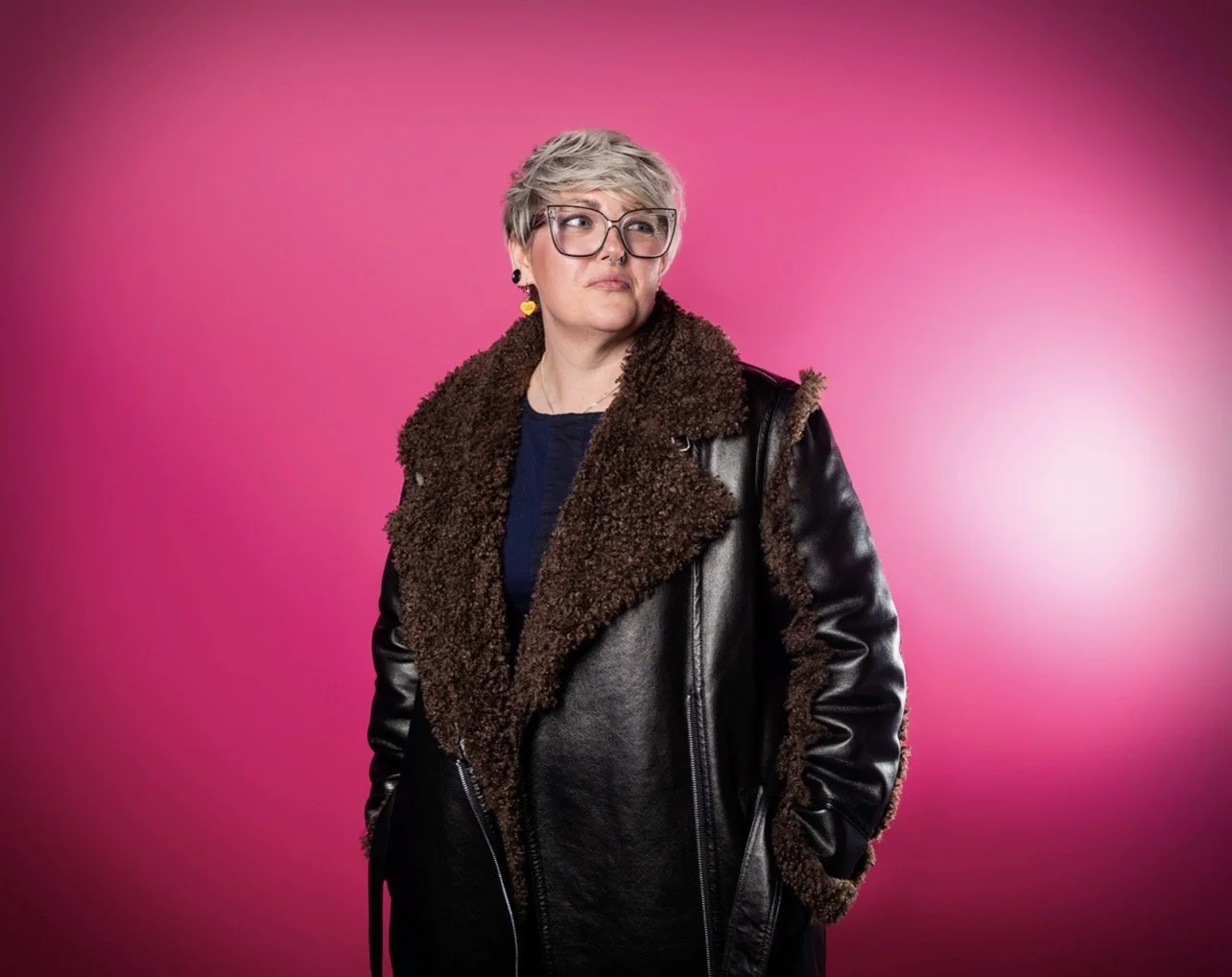 Ria Jackson with short silver hair, glasses, and earrings standing against a pink background, wearing a black leather jacket with a brown shearling collar. Photography by Haydn Brown