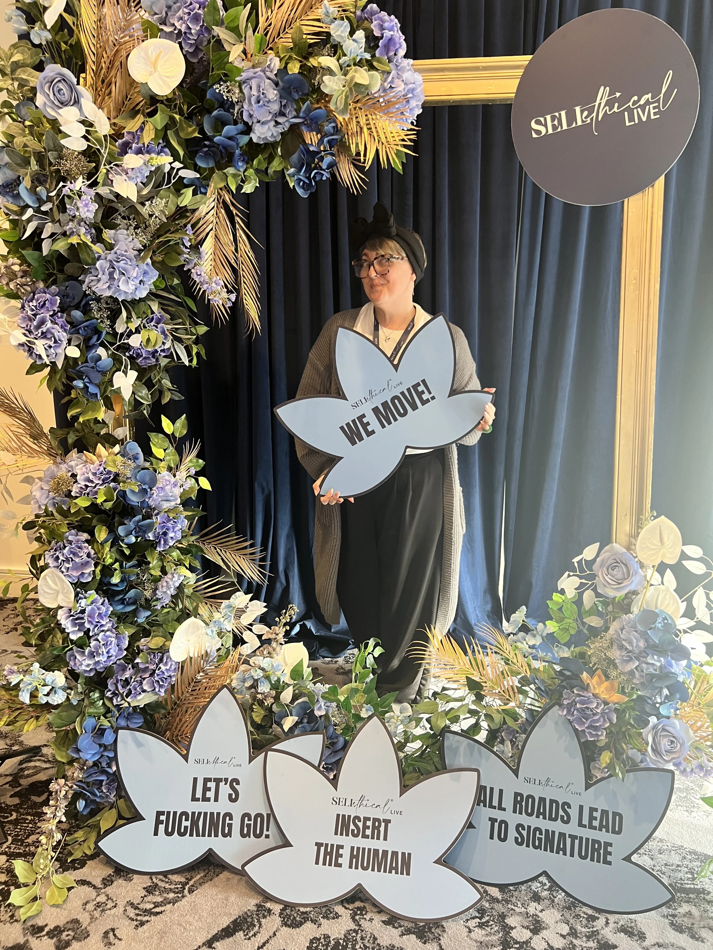A woman standing in front of a decorative floral backdrop holding a speech bubble sign with the text "WE MOVE!" at an event called 'SELF/ethical LIVE'. The backdrop features purple and blue flowers with green leaves, and several other speech bubble signs with humorous or motivational phrases are arranged at the bottom.