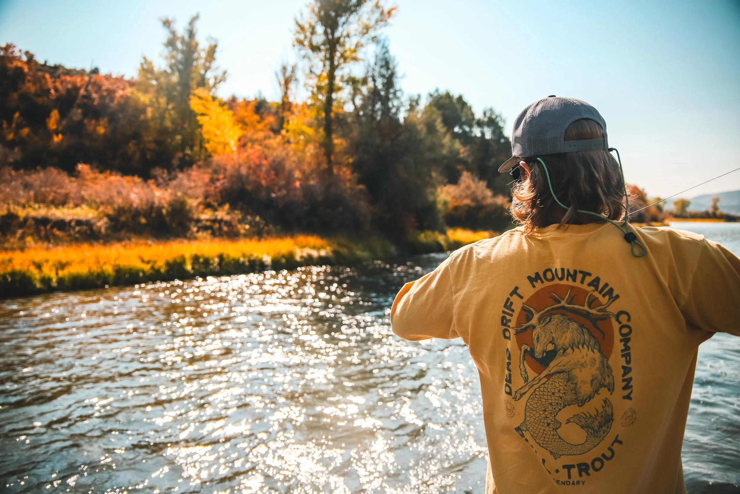 A person with long hair wearing a gray hat and sunglasses, facing away, standing by a river with trees displaying fall colors in the background, during bright sunlight.