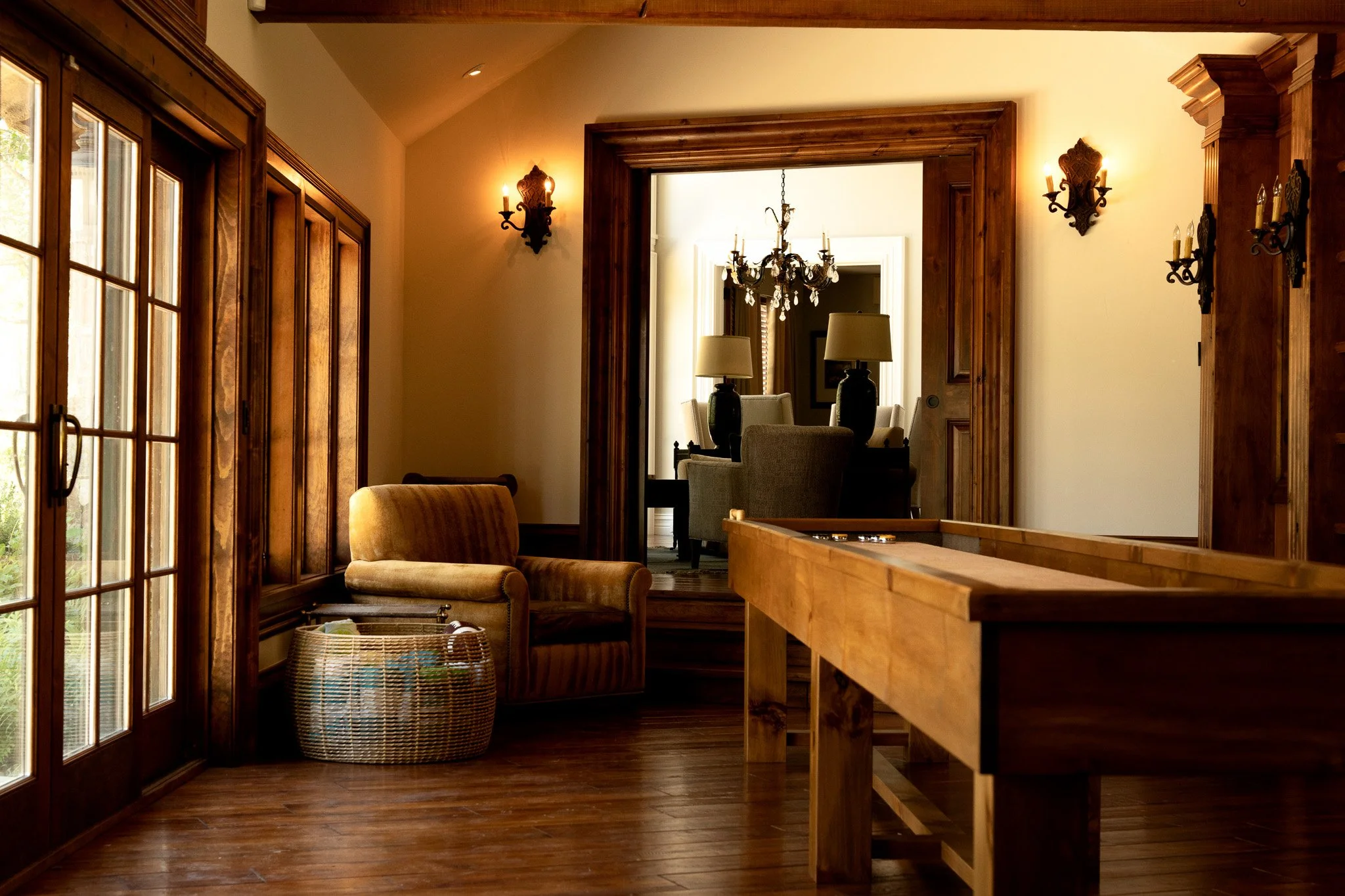 A cozy living room with wooden trim and flooring, featuring a cushioned armchair, a basket, large windows, and a view into a dining area with a chandelier, candles, and beige chairs.