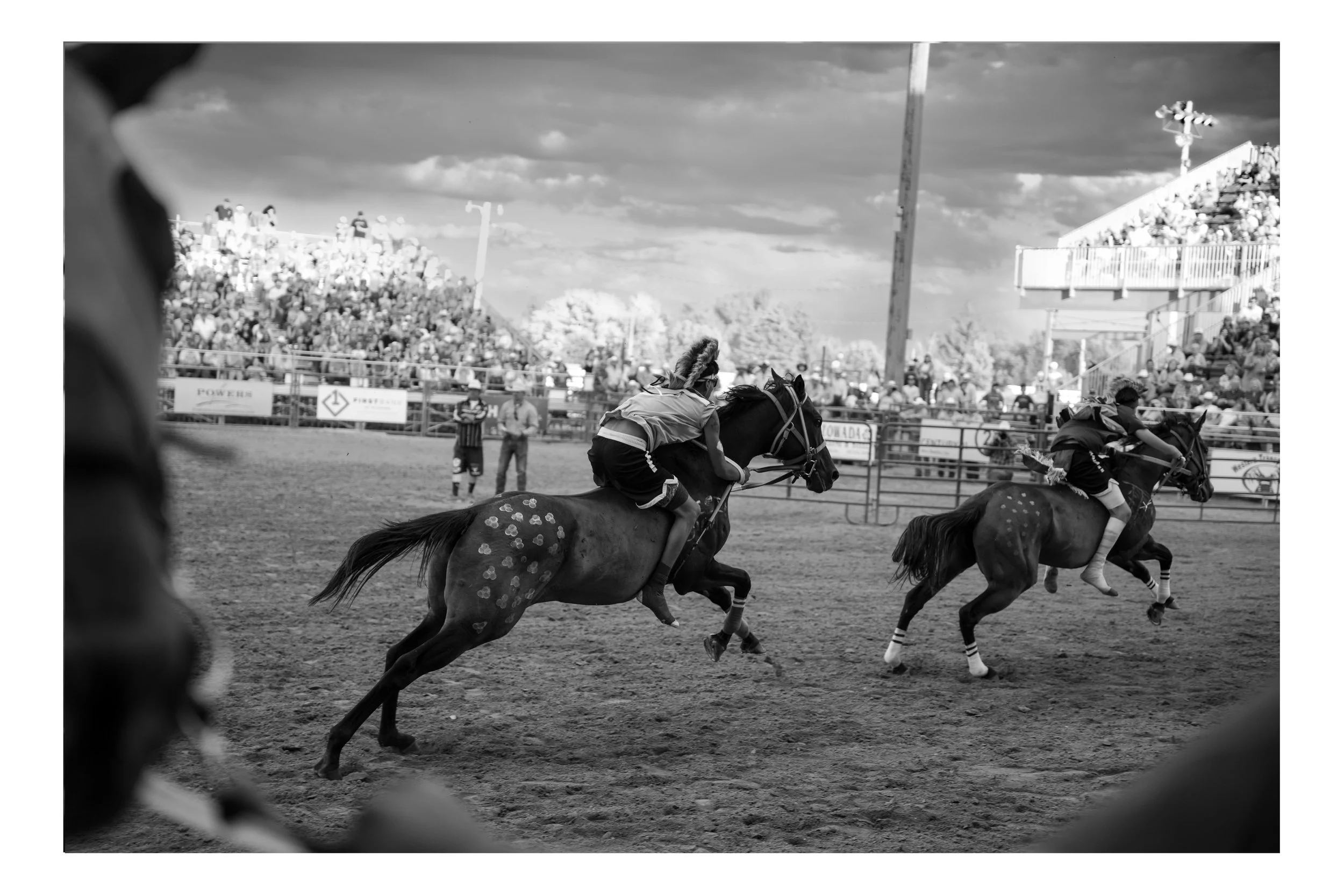 A black and white photo of a rodeo event with horse riders in action, a large crowd in the stands, and a cloudy sky overhead.