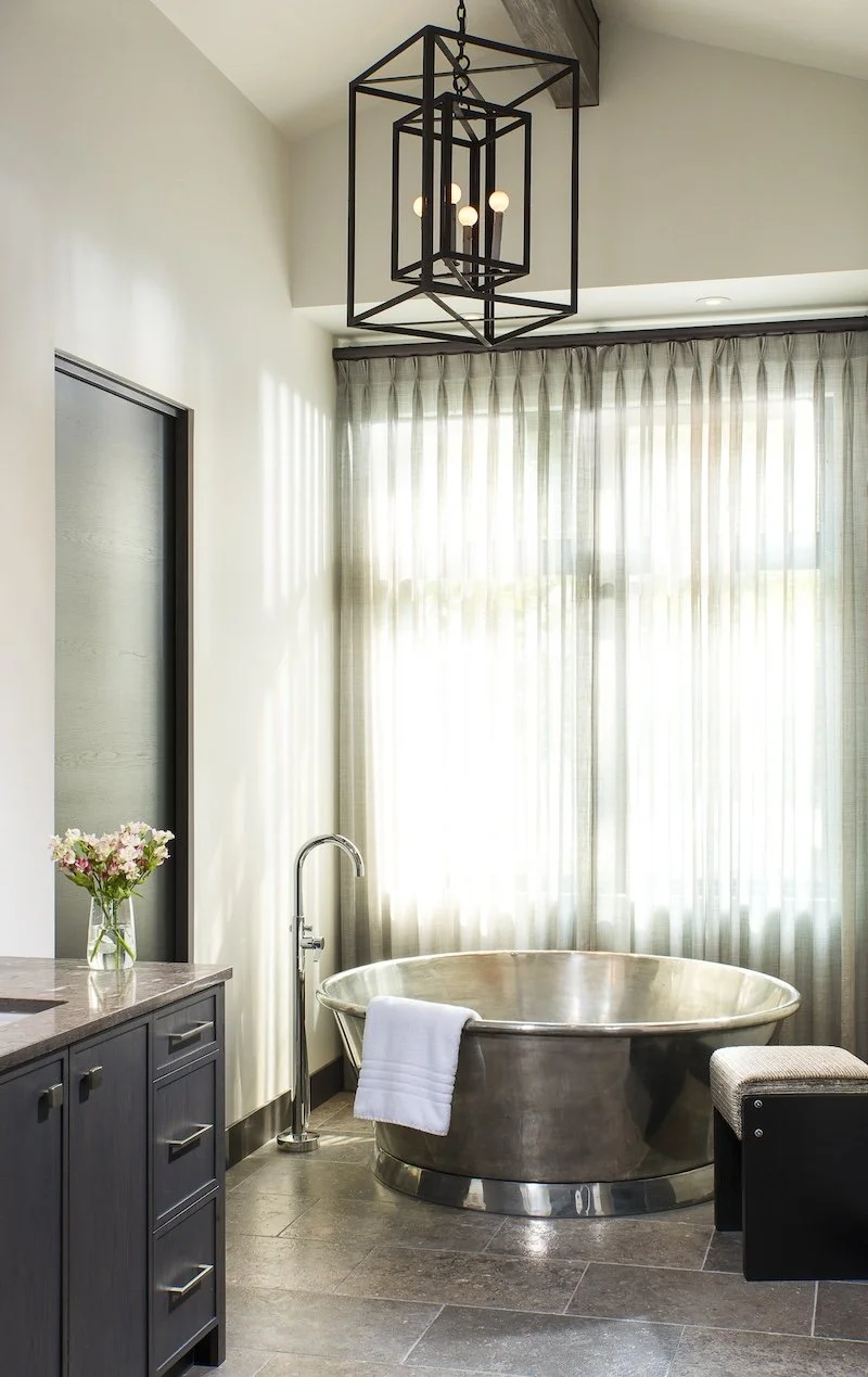 A modern bathroom with a freestanding metallic bathtub near large windows with sheer curtains, a dark cabinet with a flower vase, and a geometric black chandelier hanging from the ceiling.