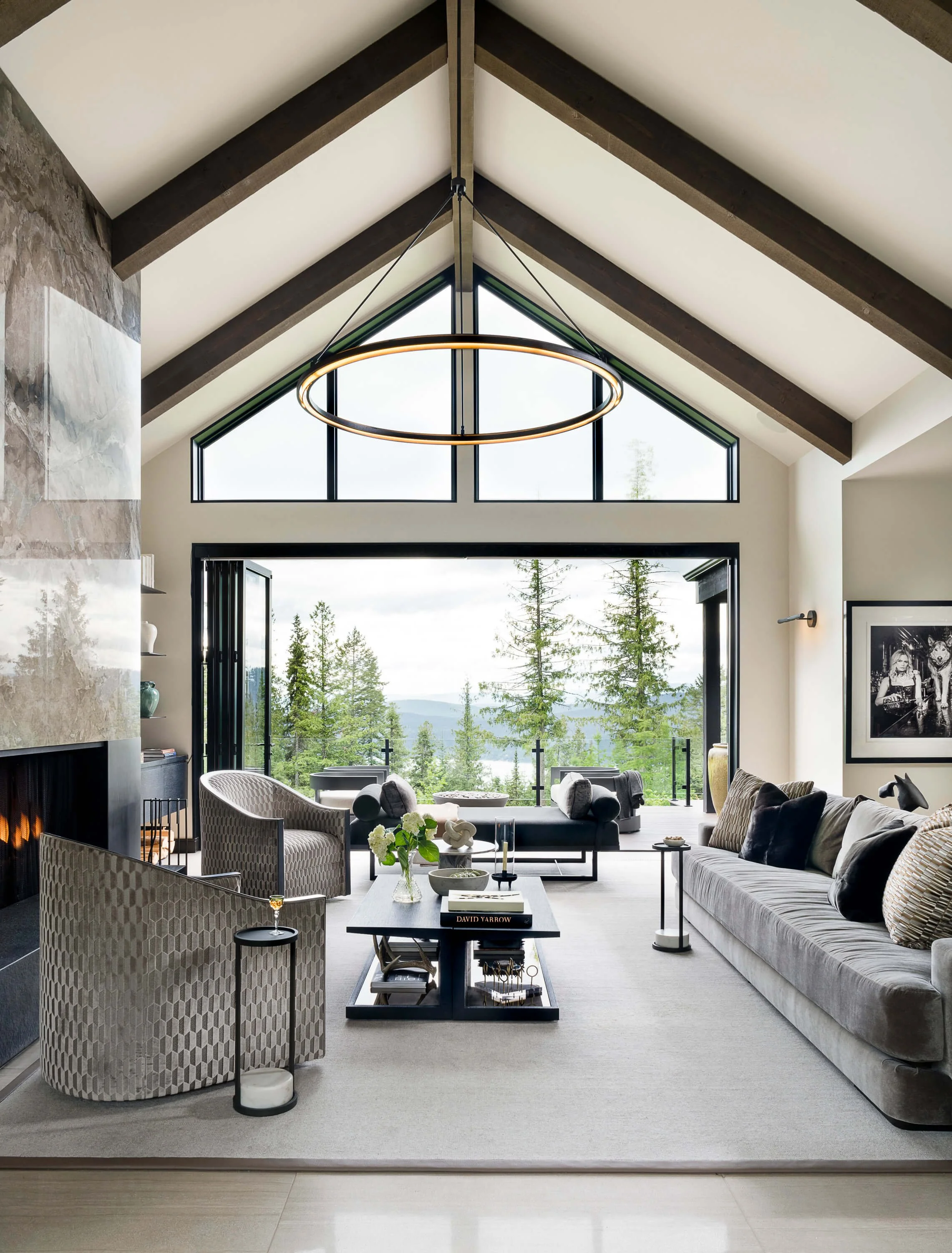 Modern living room with large window overlooking trees, featuring gray sofas, patterned armchairs, a black coffee table, and mid-century modern decor.