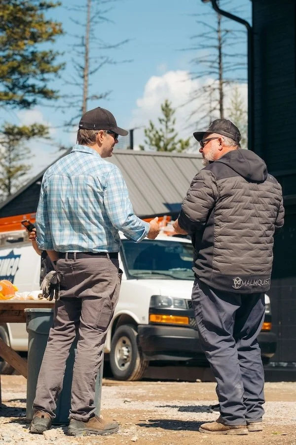 Two men stand outdoors talking near a white van, with one holding a phone. The man on the left wears a baseball cap, plaid shirt, and cargo pants, while the man on the right wears a cap, black jacket, and dark pants. There are trees and a building in the background.
