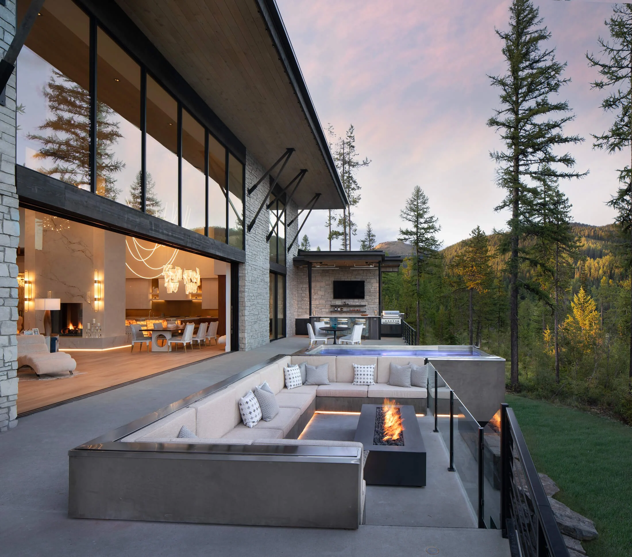 Modern house with large glass windows and outdoor living area surrounded by tall trees under a cloudy sky during sunset.